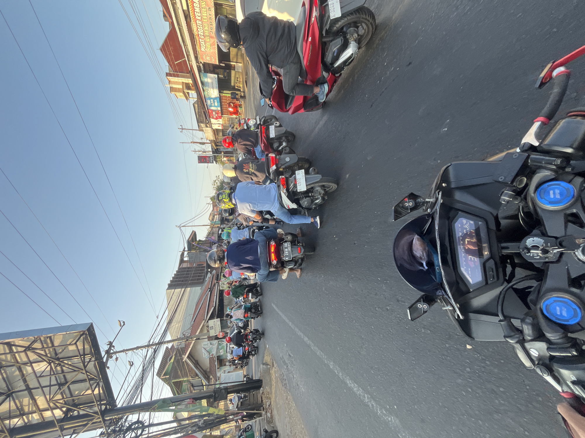 This is a first-person perspective, eye-level, medium shot photograph taken on a street in Kalinyamatan, Indonesia. The scene captures a busy traffic flow of motorcycles on a paved road. The sky is clear and blue, suggesting it is daytime. The sun is casting a warm light on the buildings lining the street, which appear to be shops or commercial establishments with awnings and signs. Electrical wires are strung across poles overhead.
In the foreground, a motorcycle's dashboard is visible, displaying speed and time. Several other motorcycles are moving in the same direction as the photographer, with drivers and passengers wearing helmets and casual clothing. Some of the motorcycles are red, while others are white or black. The license plates on some of the motorcycles are partially visible, with alphanumeric characters.
The traffic appears to be moderate, with motorcycles in multiple lanes. The road surface is asphalt with white lane markings. The overall impression is one of a typical urban or semi-urban environment in Indonesia during daylight hours, with a focus on the prevalent mode of transportation being motorcycles.