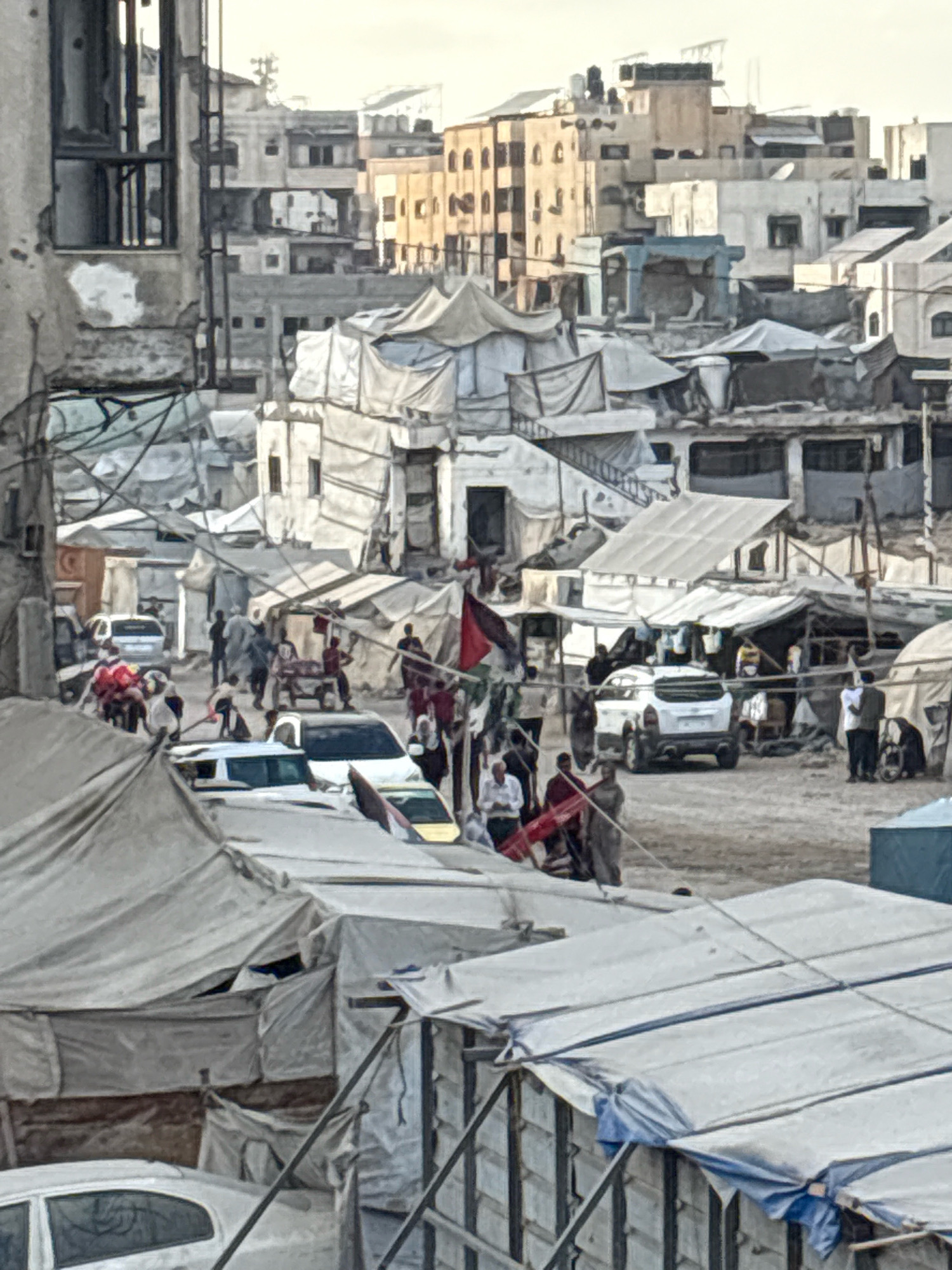 The image depicts an outdoor scene in Gaza City, Palestinian Territories, characterized by a densely populated area with makeshift structures. In the foreground, a close-up view of a temporary shelter with a blue tarpaulin roof covers what appears to be a structure built from corrugated metal. Behind this, a series of white tents and structures are visible, many with tarpaulins used as roofing.
Further into the scene, several white vehicles, including a hatchback and an SUV, are parked on a dirt or gravel surface. People are present throughout the image, some gathered in small groups, others walking. A group of individuals appears to be interacting around a table or cart. One person is holding a Palestinian flag.
In the background, a mix of damaged and intact buildings are visible, suggesting a war-torn or heavily affected urban environment. The structures range from multi-story buildings to smaller, more informal constructions. The overall impression is one of a community living in temporary or damaged housing amidst a cityscape. The lighting suggests it is daytime, possibly late afternoon due to the soft light.