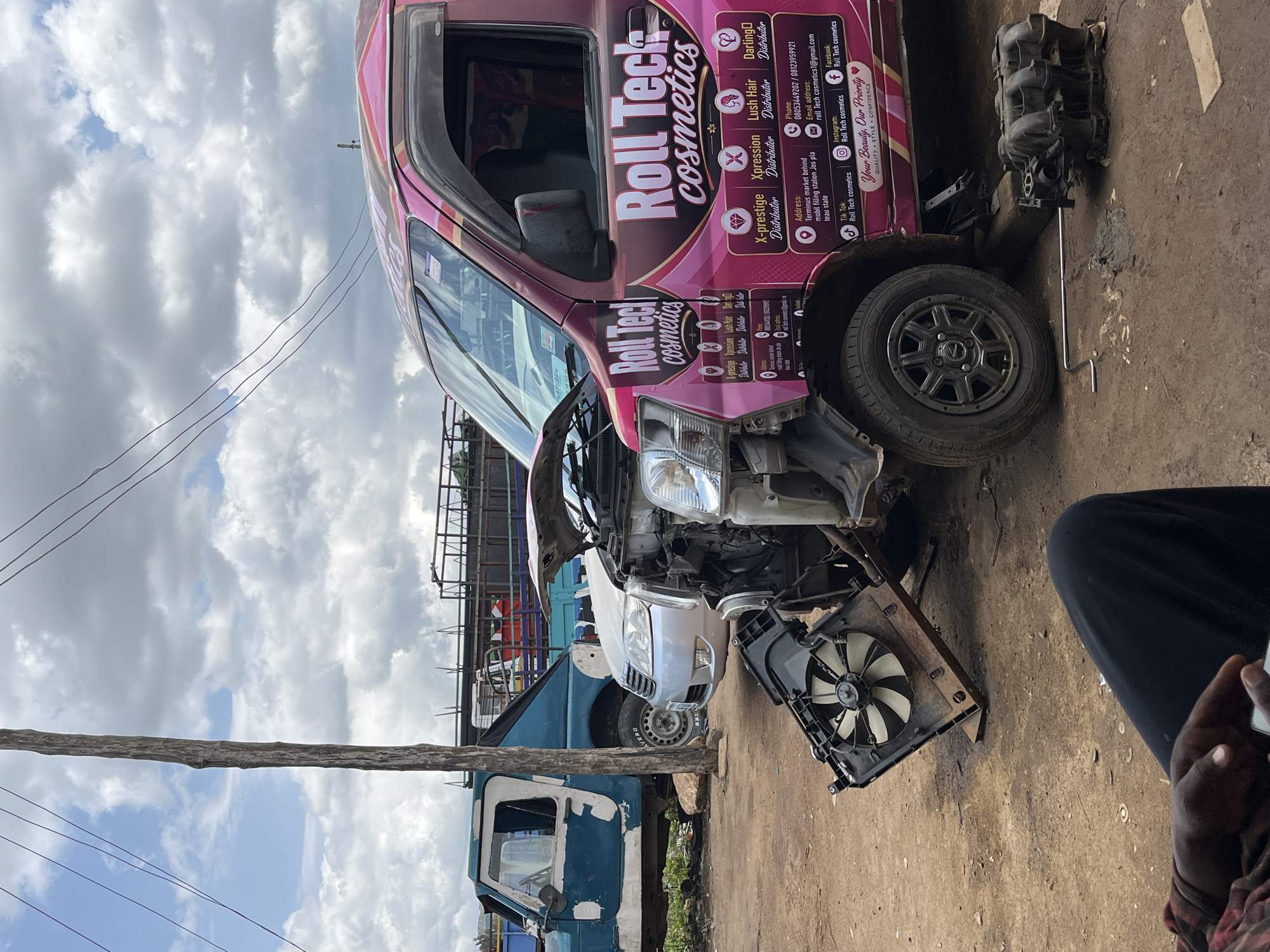 The image depicts a chaotic scene outside what appears to be a mechanics workshop or scrapyard in Rafin Par, Nigeria. In the foreground, a person wearing a red and black checkered shirt and black pants is seated, holding a smartphone. In the middle ground, a pink van, branded "Roll Tech cosmetics" with various distributors listed, has its hood open and appears to be undergoing repairs. A car fan and radiator are detached and placed on the ground in front of the van. To the left, a turquoise and white jeep is parked, and further behind it, a large truck with a metal cage is visible. The ground is uneven dirt, and debris is scattered around. The sky is overcast with scattered clouds. The overall environment suggests a busy, working area with vehicles in various states of repair or disuse.