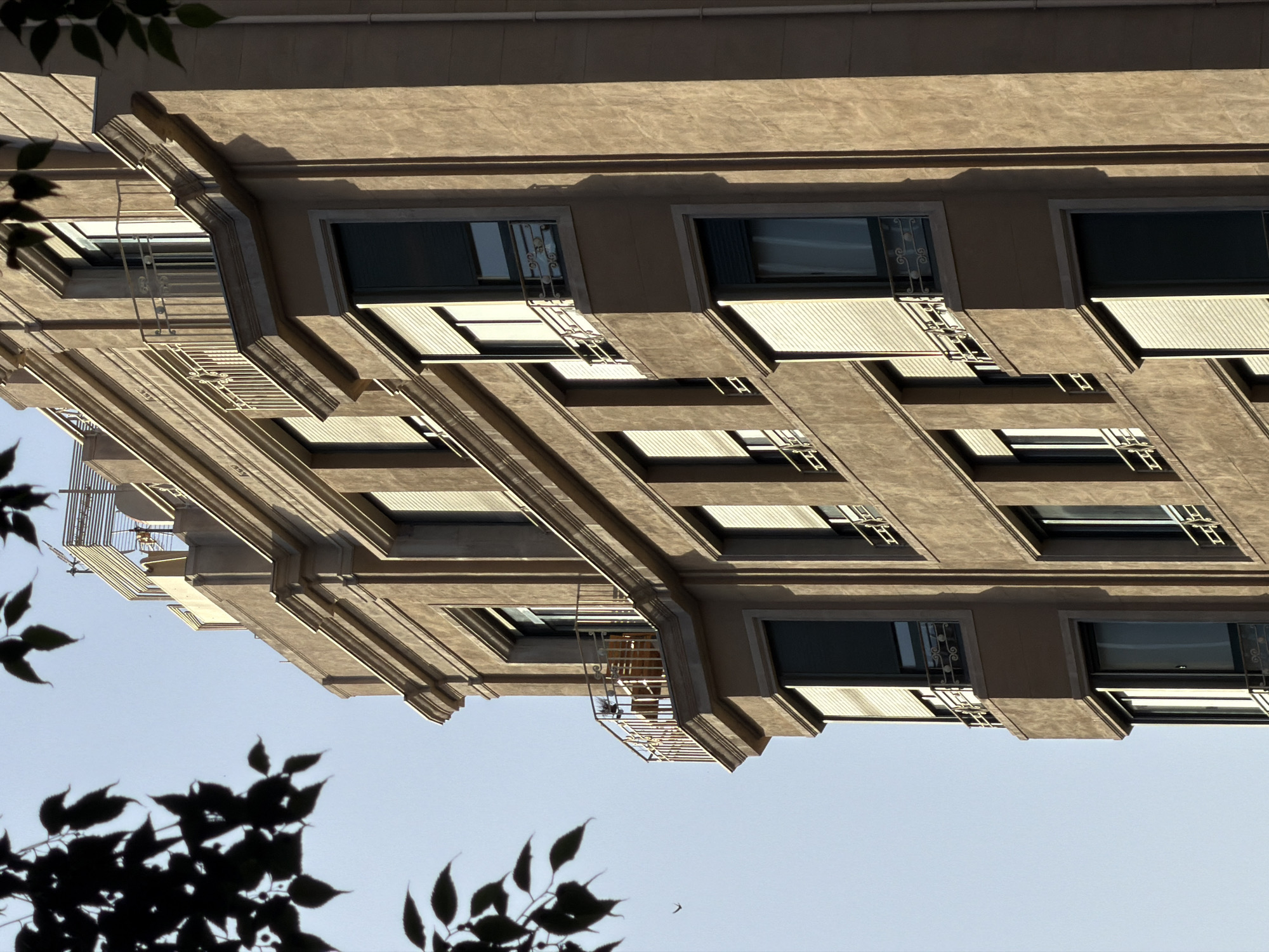 The image shows a detailed close-up of a tan-colored, multi-story building with a distinct architectural style. The building's facade features several windows, some with closed roll-down shutters and others revealing a glimpse of the interior. Balconies with decorative metal railings are present on various levels. The building is made of stucco or a similar textured material. The top of the building shows a flat roof with some antenna-like structures and railings. In the foreground, dark green leaves and branches of a tree are partially obscuring the view of the building, creating a silhouetted effect against a clear, pale blue sky. The lighting suggests it is daytime, with shadows cast by the building's architectural features and the leaves. There are no people visible in the image. The style of the building, particularly the balconies and decorative elements, is suggestive of early 20th-century architecture common in Barcelona.