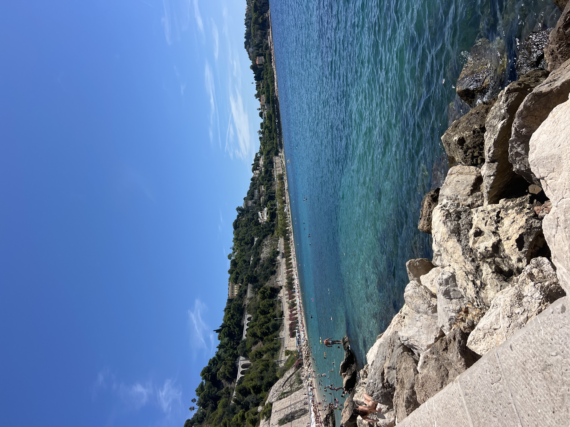The image depicts a vibrant seaside scene in Villefranche-sur-Mer, France, on a clear, sunny day. In the foreground, large, light-colored rocks line the edge of the water, with a paved walkway visible on the left. The turquoise water of the Mediterranean Sea is crystal clear, revealing the rocky seabed.
The beach, situated along a crescent-shaped bay, is populated with numerous sunbathers. Many people are seen relaxing on the sand, swimming, and wading in the shallow waters. The beach is backed by a lush, green hillside, dotted with buildings and trees, creating a picturesque backdrop. A road with a retaining wall separates the beach from the hillside. Several structures, including what appear to be arches and buildings, are visible ascending the slope.
In the mid-ground, a man stands on a rock at the water's edge, facing away from the camera. A woman is seated on the rocks in the lower left corner of the image, also facing away. The water is calm with gentle ripples. The sky above is a brilliant, cloudless blue, with a few wispy clouds in the distance. The overall atmosphere is one of a bustling summer day at a popular coastal destination.