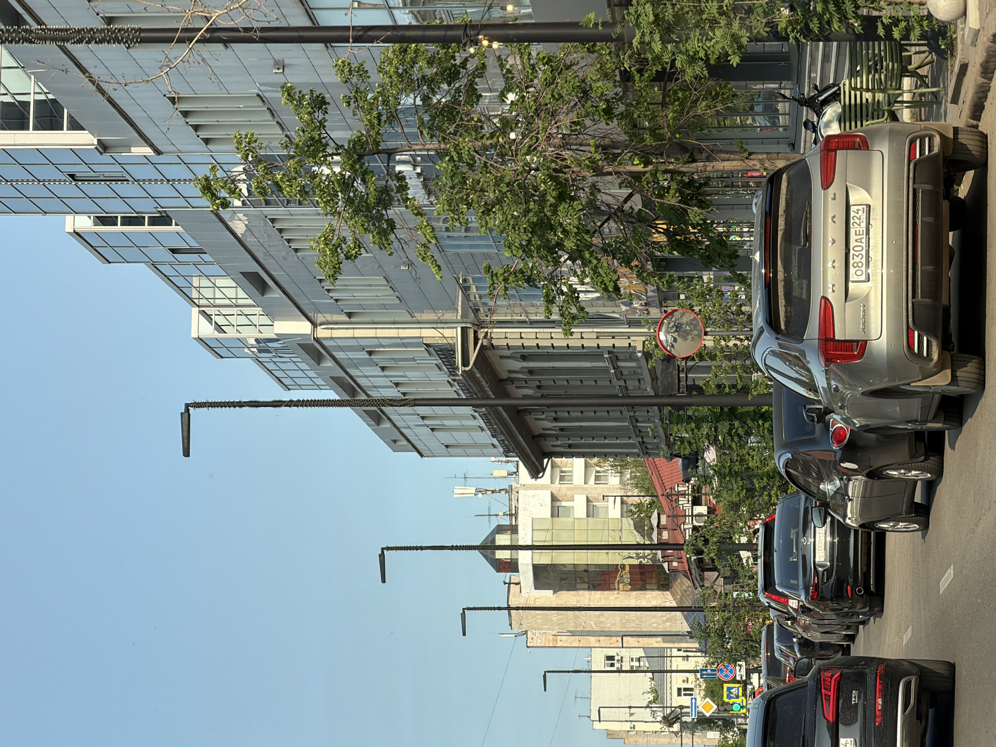 The media file shows a street scene in Krasnoyarsk, Russia, with a row of cars parked along the side of the road. The buildings on the left are modern, with glass facades and balconies. Trees with green leaves are scattered along the street, providing shade. The sky is clear and blue, suggesting a sunny day. The time of day appears to be daytime, likely late morning or early afternoon, given the angle of the sunlight.
Several cars are visible, including a silver Haval Jolion with the license plate "0830AE 224" and a dark-colored Audi SUV in the foreground. The cars are parked parallel to the sidewalk. Streetlights with a distinctive zig-zag design are spaced along the street. In the distance, more buildings can be seen, some appearing older with multiple windows and balconies, while others have a more contemporary glass-covered design. Traffic signs are visible, including a pedestrian crossing sign and a sign indicating a route. A convex mirror, often used to improve visibility at intersections, is mounted on a pole. The overall impression is a typical urban streetscape on a clear day.
