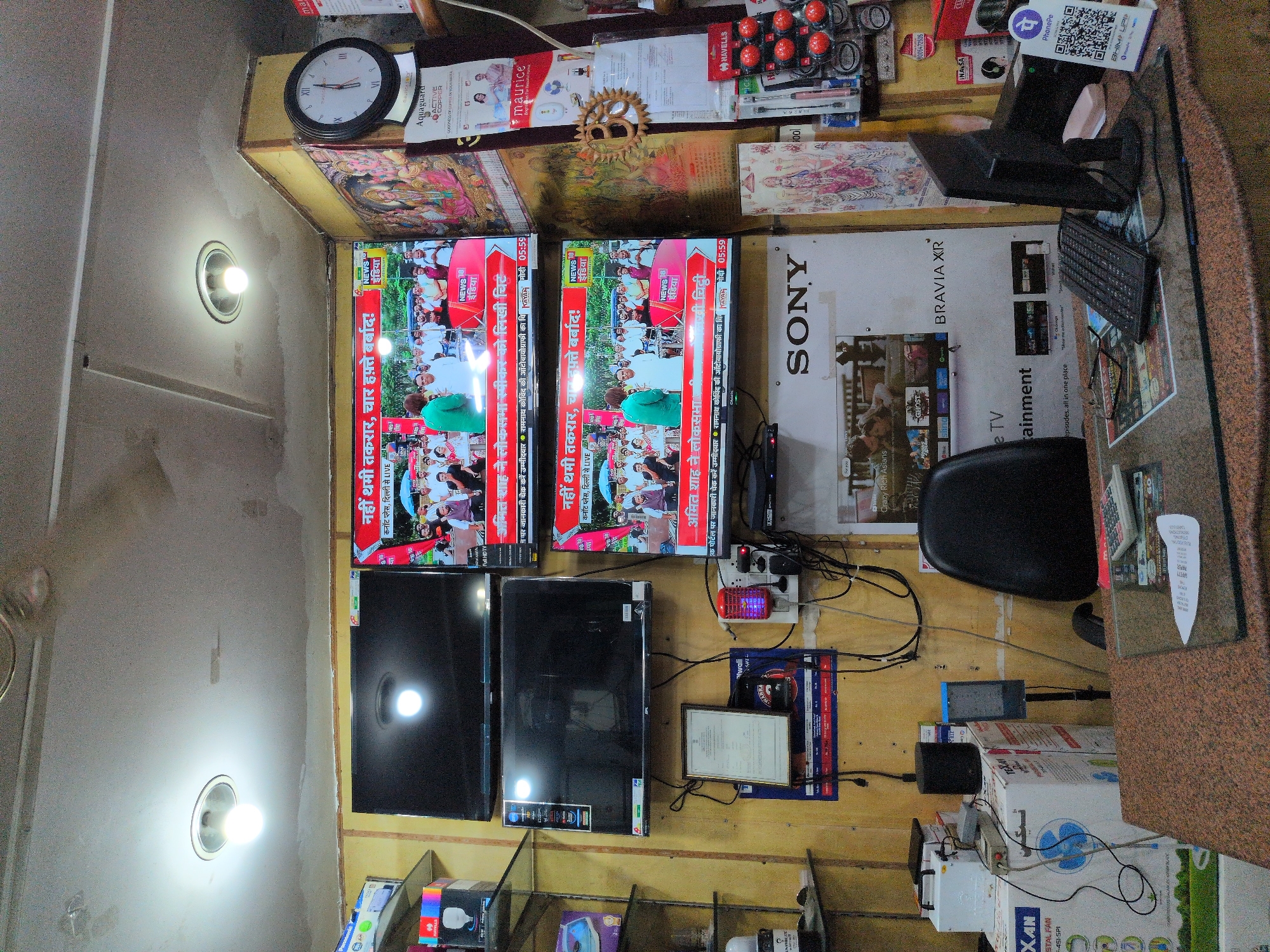 The image depicts the interior of a small electronics shop in Kullu, India, likely on a street level given the overhead lighting and display of goods. The shop is cluttered with various electronic items. Dominating the scene are three television screens displaying news channels. The top screen shows a news program with text in Hindi, and the middle and bottom screens also display news segments, featuring people and Hindi text. To the right of the televisions, a large round analog clock is mounted on a yellow wall, which is adorned with religious imagery, specifically a depiction of Hindu deities. Below the clock, several promotional posters and product advertisements are visible.
In the foreground, a counter is laden with items including a computer monitor, keyboard, eyeglasses, and a small QR code sticker for a payment app, indicating that digital transactions are accepted. Various boxes of electronics, including a fan, are stacked behind the counter. Electrical appliances, such as a mosquito repellent device, are plugged into a power strip on the wall. A ceiling fan is also visible, suggesting a warm climate. The overall impression is of a busy, functional retail space catering to local consumers.