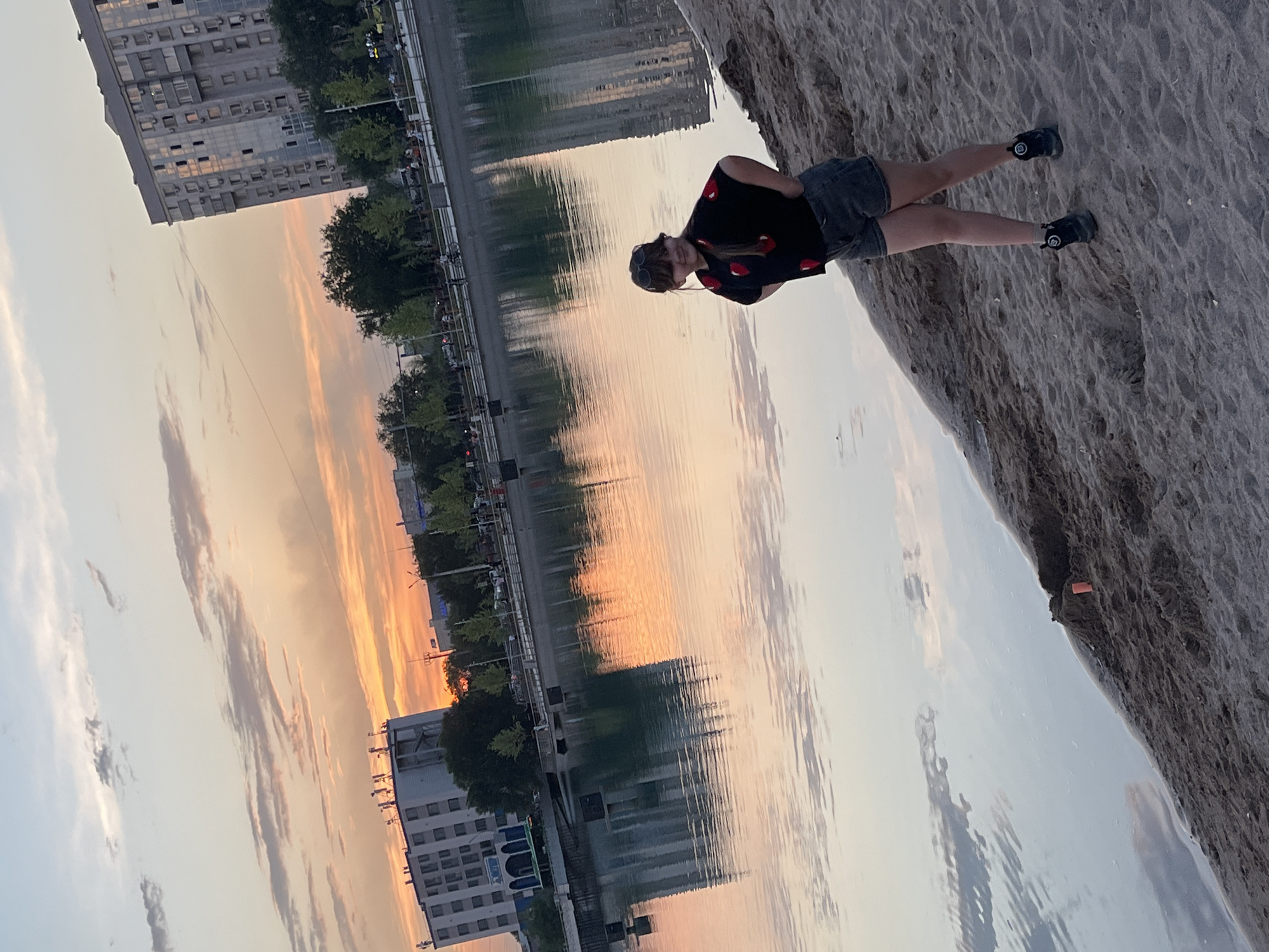 A young girl stands on a sandy embankment overlooking a body of water. The sky above is painted with the warm hues of sunset, casting a soft glow on the scene. In the background, apartment buildings line the opposite shore, their reflections shimmering on the water's surface. The girl is wearing a black t-shirt with red strawberry prints and denim shorts. She has sunglasses perched on her head and is looking to her right. The environment appears to be a recreational area within the city, possibly a park or a lakeside promenade. The lighting and reflections suggest a peaceful evening setting. This scene is likely from the city of Almaty, Kazakhstan, given the architectural style of the buildings and the presence of a body of water often found in urban parks.