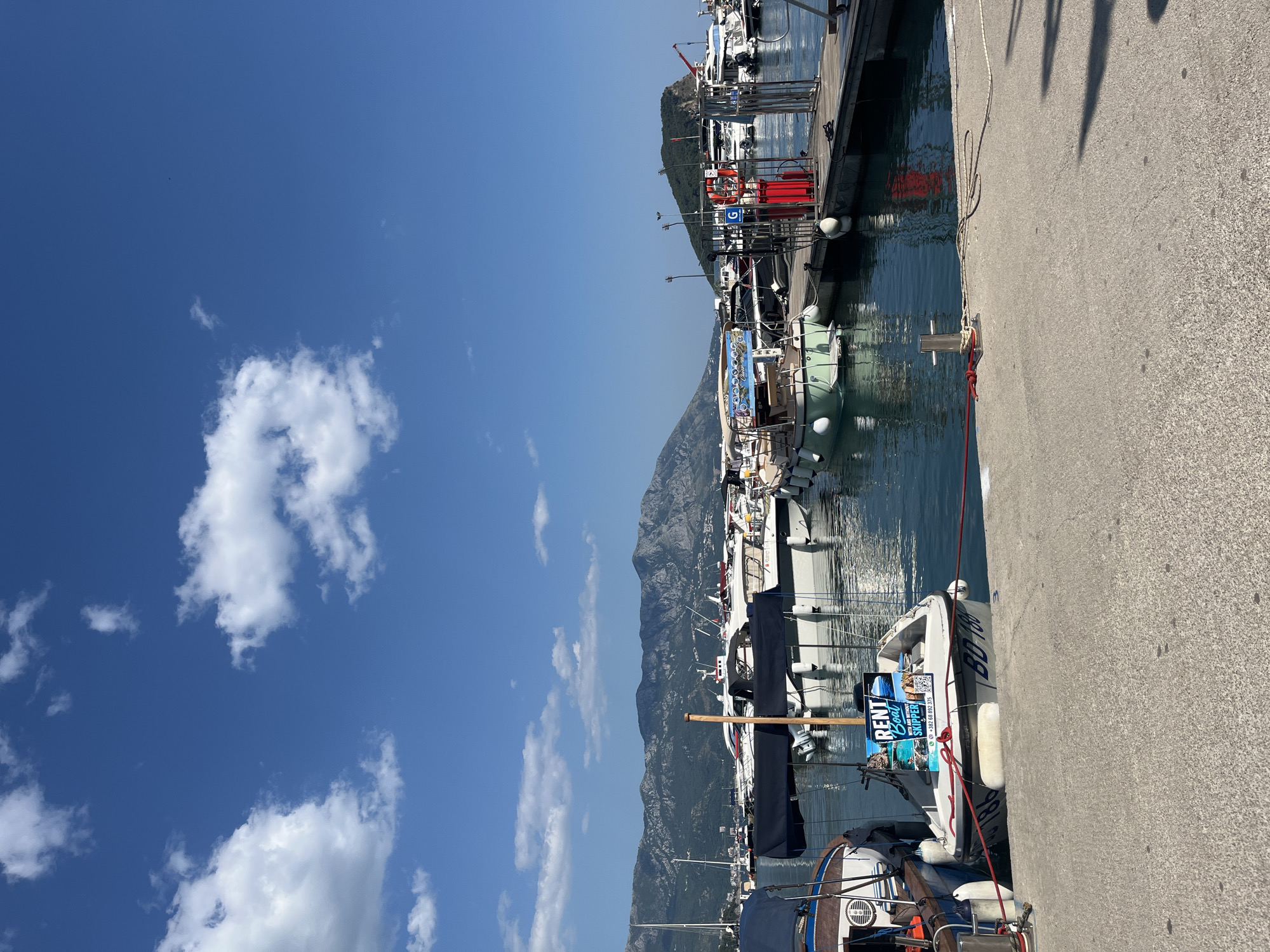 The image depicts a marina in Budva, Montenegro, on a clear, sunny day. The foreground shows a concrete pier with various boats docked. A small white boat with a "RENT Boat" sign and phone number is prominently displayed. Several other boats, including larger yachts and a green boat, are moored alongside. In the middle ground, a wooden pier with red fuel pumps and safety equipment is visible. The background features a panoramic view of mountains covered in lush green vegetation, with a small island topped by a church and a red crane in the distance. The sky is a vibrant blue with scattered white clouds. The overall scene is one of a bustling harbor on a picturesque Mediterranean coastline.