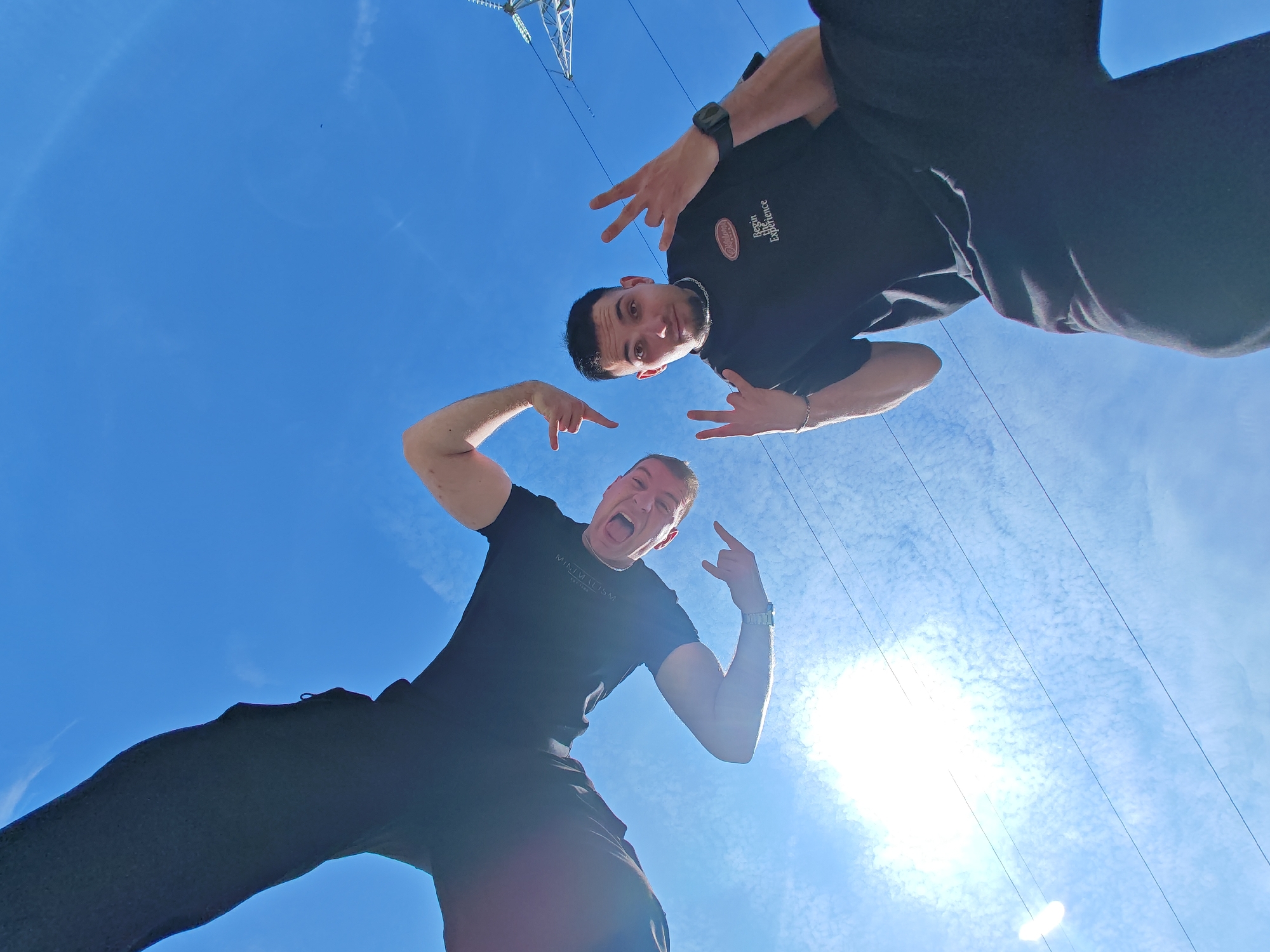A low-angle photograph features two adult males posing against a clear blue sky with scattered clouds, multiple power lines, and the upper section of a metal electricity pylon visible in the upper-left quadrant. The individual on the left, positioned closer to the camera, wears a black t-shirt with white "MINIMALISM EST. 1989" text and black pants. He has short light hair, a silver-toned watch on his left wrist, and displays an open-mouthed expression as if yelling. His right hand is in a "rock on" gesture, and his left index finger points downwards. The individual on the right, positioned slightly higher, wears a black t-shirt with an orange and white circular "Begin The Experience" logo and black pants. He has short dark hair and a goatee, looks directly at the camera with a subtle smile, and holds both hands in "peace signs." A black smartwatch is on his left wrist, and a silver chain necklace is visible. A bright sun flare emanates from the lower right portion of the image. The scene is located in Shumen, Bulgaria, in an outdoor area characterized by power transmission infrastructure, suggesting a location on the city's outskirts or within an industrial zone.