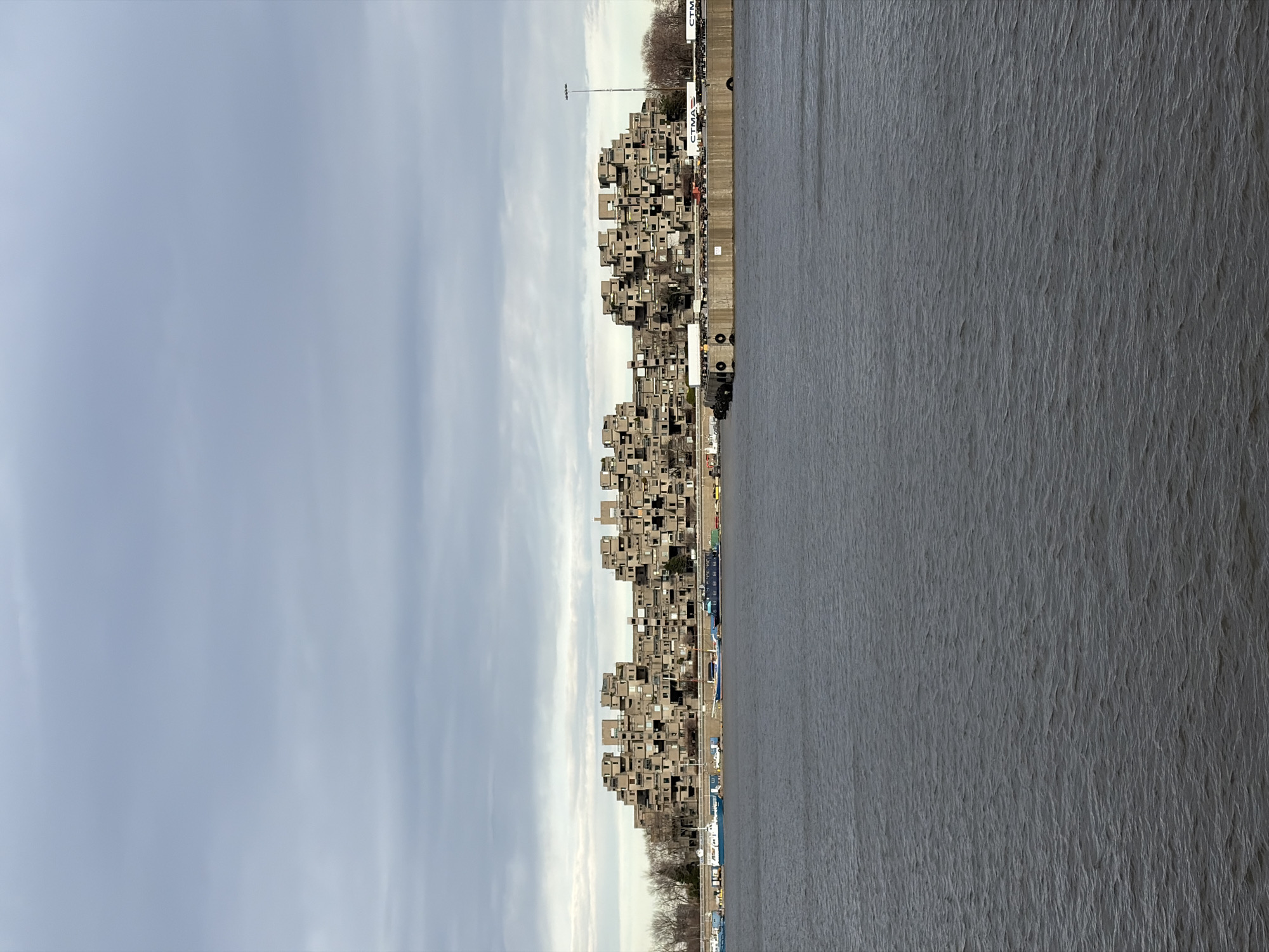 The image shows Habitat 67, a modular housing complex in Montreal, Canada, built for Expo 67. The building is situated next to a body of water, presumably the Saint Lawrence River, with a cloudy sky above. The architecture is characterized by stacked, concrete blocks, creating a distinctive, cubist-like structure. A road and other infrastructure elements run alongside the building.