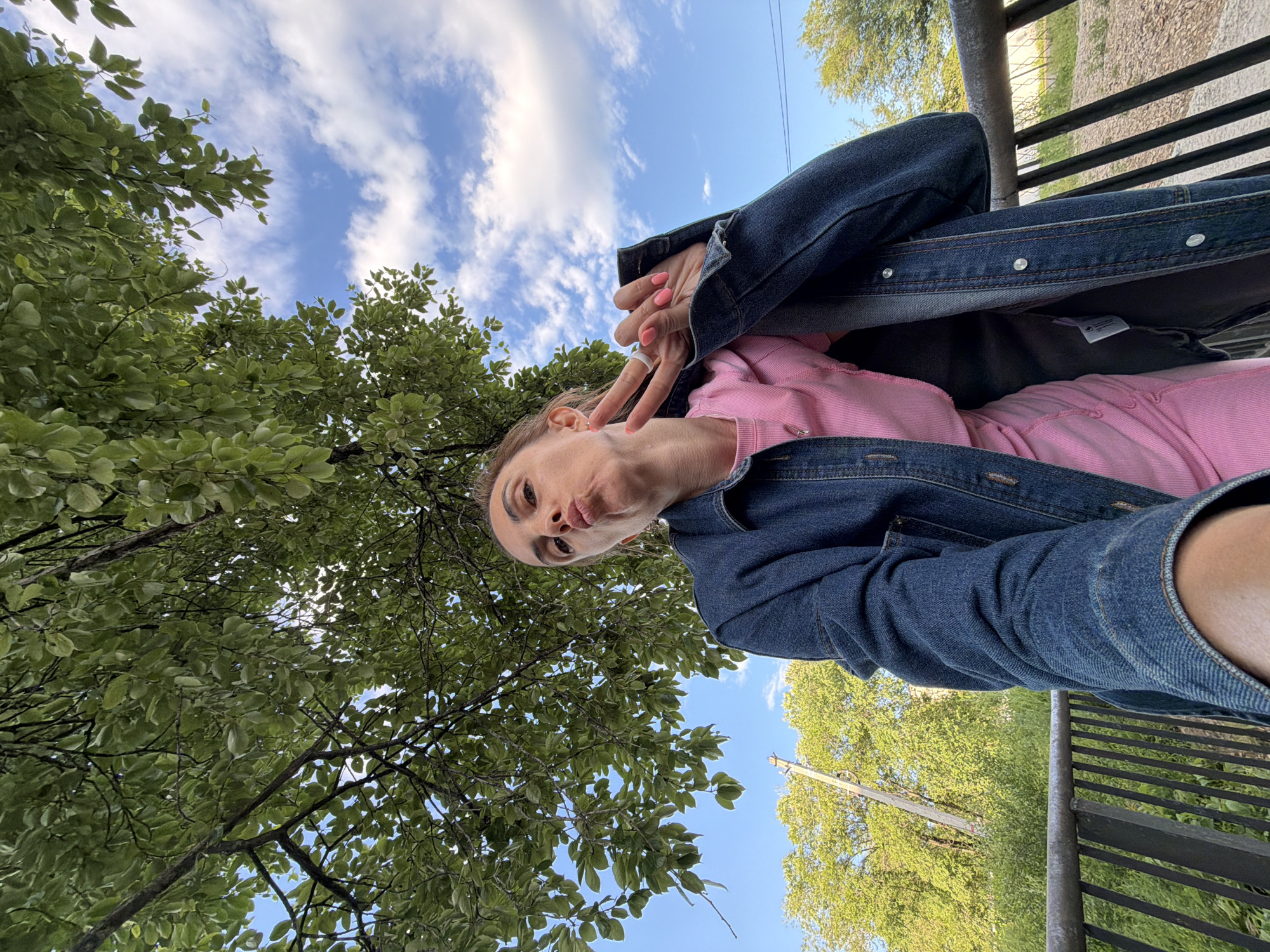 A woman is captured from a low angle, looking up towards the camera with a puckered expression and her fingers forming a peace sign. She is wearing a pink top and a denim jacket. The background consists of a tree canopy overhead, a blue sky with white clouds, and lush green foliage and trees. A metal railing is visible in the foreground. The image appears to have been taken outdoors, potentially in a park or a natural setting within the city of Birobidzhan, Russia, given the presence of mature trees and what seems to be a somewhat rural or park-like environment.