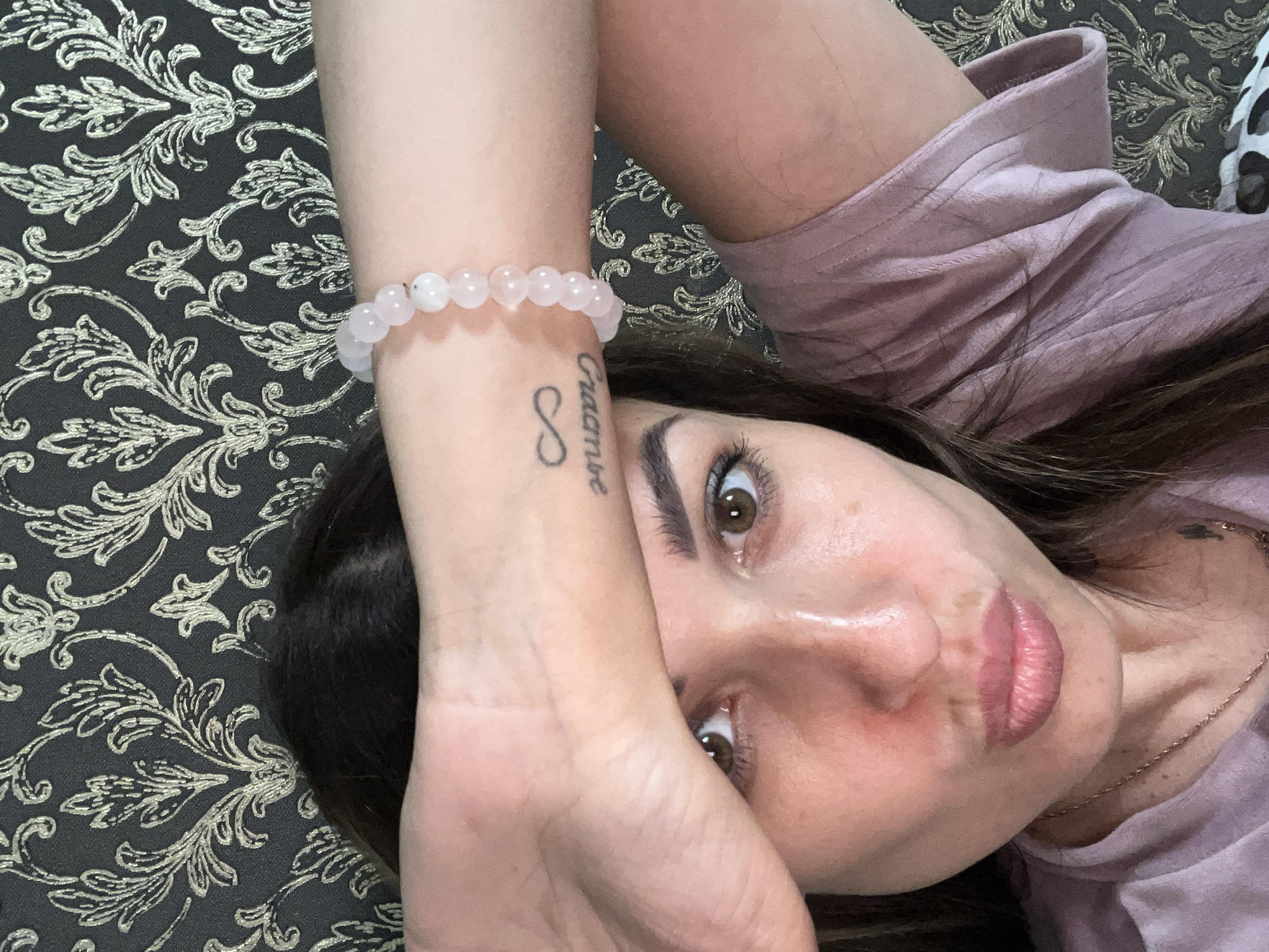A close-up, overhead selfie shows a woman with dark hair and brown eyes, her left arm raised to cover her forehead, obscuring the top of her head. She wears a pink, ribbed t-shirt and a pink beaded bracelet on her left wrist. A small infinity tattoo is visible on her wrist. Her lips are slightly pursed, and her right cheek is flushed. The background features a dark, patterned wallpaper with a gold floral design. The image appears to be taken indoors, likely in a domestic setting.