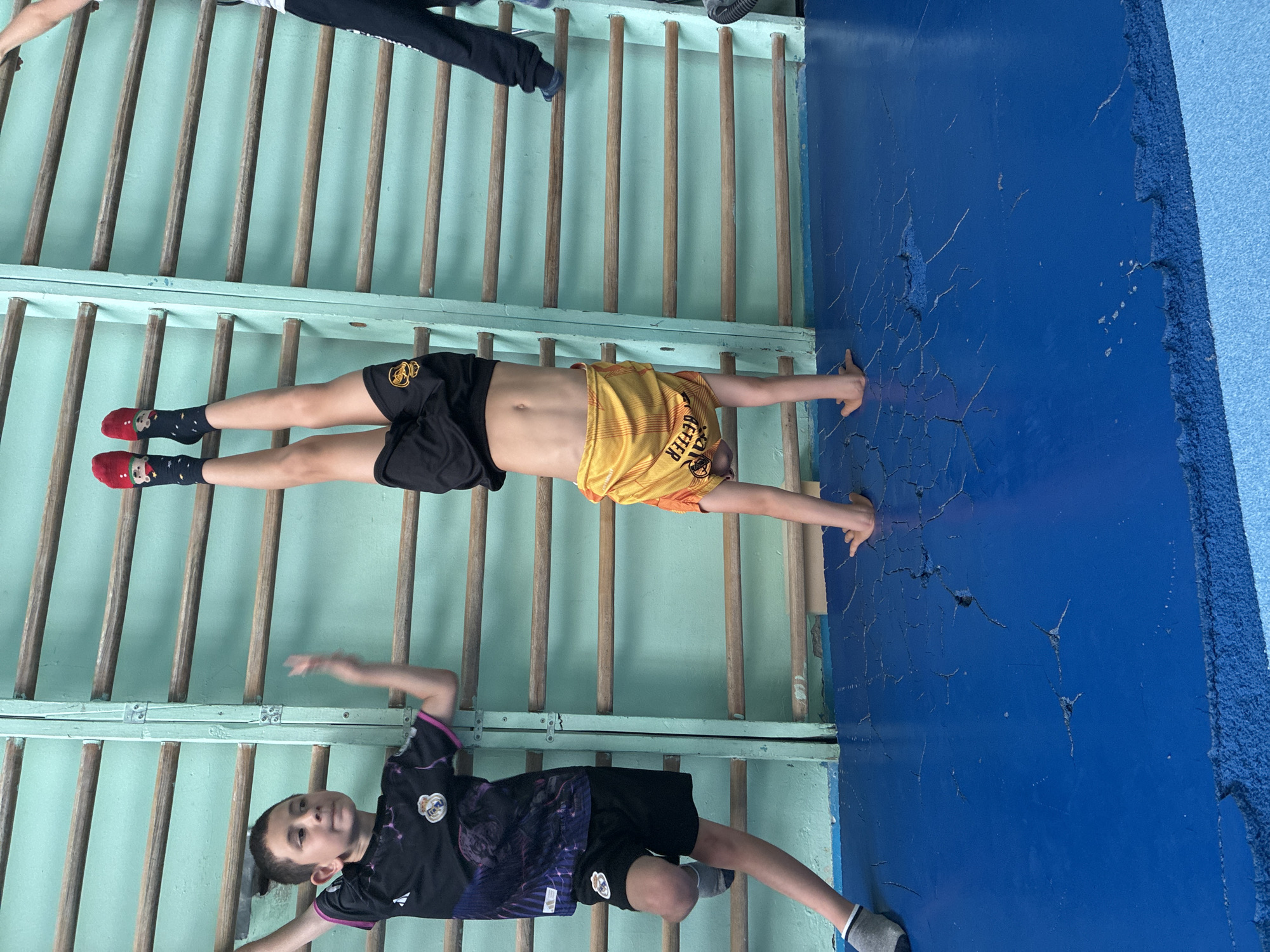 Three young boys are engaged in physical activity within what appears to be an indoor gymnasium or sports hall. The primary focus is on a wall-mounted wooden gymnastics ladder (stall bars) against a light blue-green wall.
Two of the boys are actively using the ladder. The boy at the top of the frame (if oriented correctly) is hanging from an upper rung by his hands, with his legs extended straight down. He is wearing black athletic shorts and a yellow and orange sleeveless top, on which the word "BETTER" is partially visible. The middle boy is also hanging from a lower rung, grasping it with both hands above his head. He wears black athletic shorts and distinctive red socks with a white cartoon bear design, with blue and white striped socks layered underneath.
The third boy is positioned at the bottom of the ladder, facing the camera. He is standing or leaning against the lower rungs, appearing to be resting or observing. He is dressed in a dark (black and possibly purple/blue) athletic t-shirt featuring a circular crest, similar to a football club logo, black athletic shorts, and gray and white striped socks.
To the right of the wooden ladder, there is a dark blue wall or padded surface that shows significant signs of wear, including numerous cracks and peeling paint, suggesting heavy use or age. The overall lighting indicates an indoor daytime setting. The scene captures a moment of physical training or exercise.