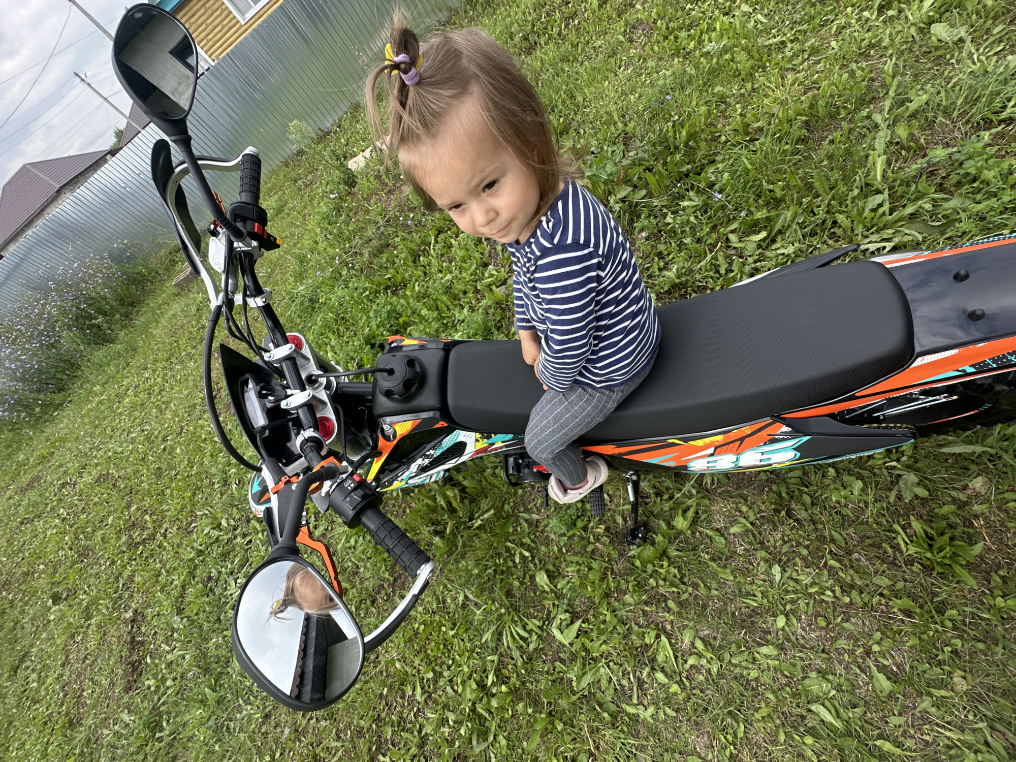 A young Caucasian girl with blonde hair in a ponytail is sitting on the seat of a dirt bike parked on a grassy area. The girl is wearing a striped long-sleeved shirt and grey pants. The dirt bike is orange, black, and white with various decals. A side mirror is visible on the handlebars, reflecting the girl's head and shoulders. In the background, there is a metal fence, a house, and green vegetation under a cloudy sky. The location is specified as Kharnavy, Russia.