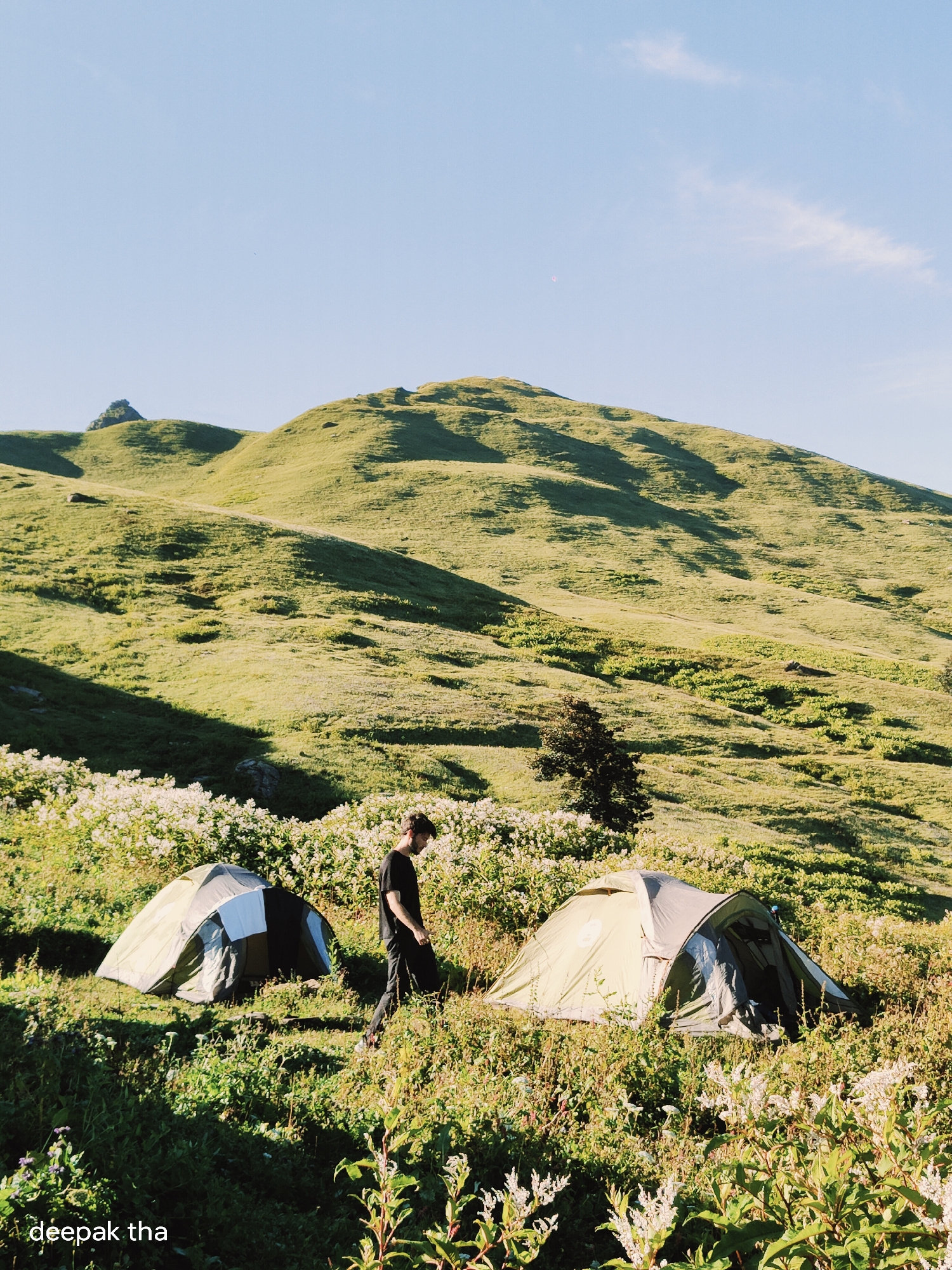 A man in a black t-shirt and dark pants walks past two tents set up in a grassy, mountainous landscape. The tents are positioned on a slope dotted with white wildflowers. In the background, rolling green hills dominate the scene under a clear blue sky. The image is labeled with "deepak tha". The location is in the city of Manali, India.