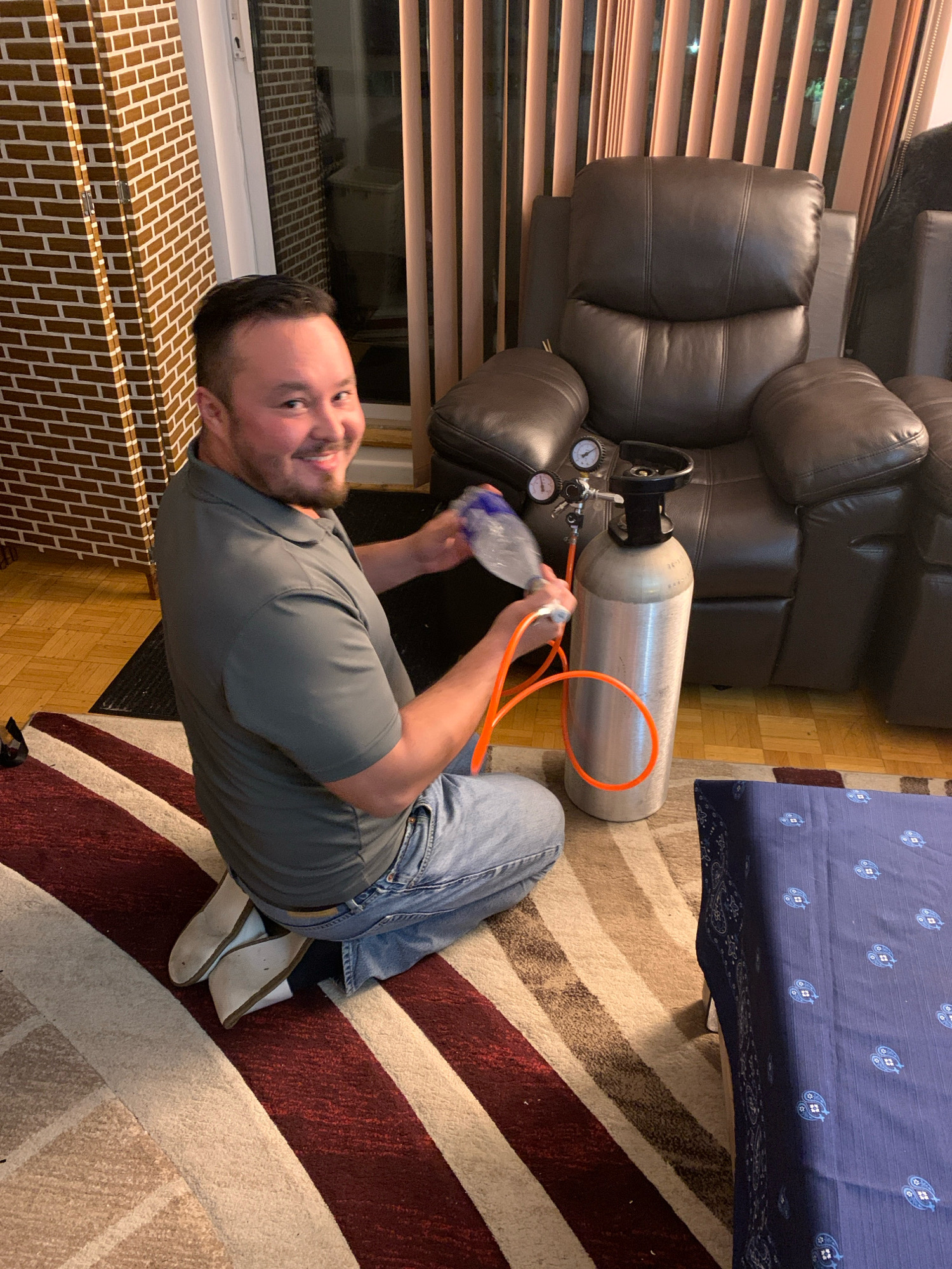 A man kneels on a rug in a room, holding a plastic water bottle attached to a gas cylinder by an orange hose. The man is wearing a grey polo shirt and blue jeans. Behind him is a large, dark brown leather recliner chair. To his left is a tall, brick-patterned partition. The floor is hardwood, partially covered by a striped rug. A blue fabric with a white paisley pattern is visible in the lower right corner. The scene appears to be indoors, likely a residential setting. There are no specific visual cues indicating the exact location within Markham, Canada.