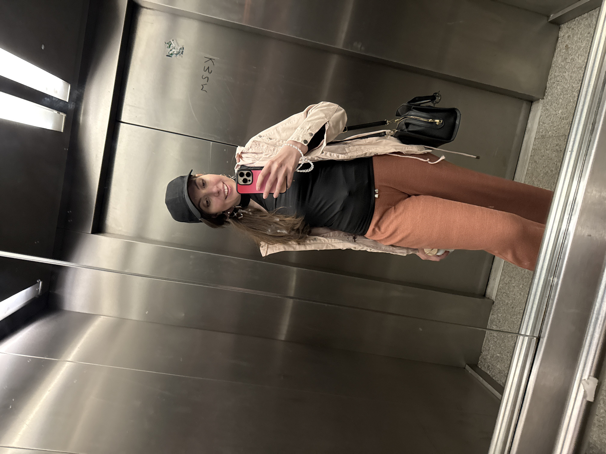 A woman is in a reflective metal elevator, taking a selfie with a pink cell phone. She is wearing a black cap, black top, light pink jacket, and reddish-brown pants. She has a black purse with a gold zipper strap over her shoulder. The elevator walls are metal with "K35M" written on one wall. Fluorescent lights are visible on the ceiling. A handrail is present in the elevator. The floor is gray. The location is Unknown, Unknown.