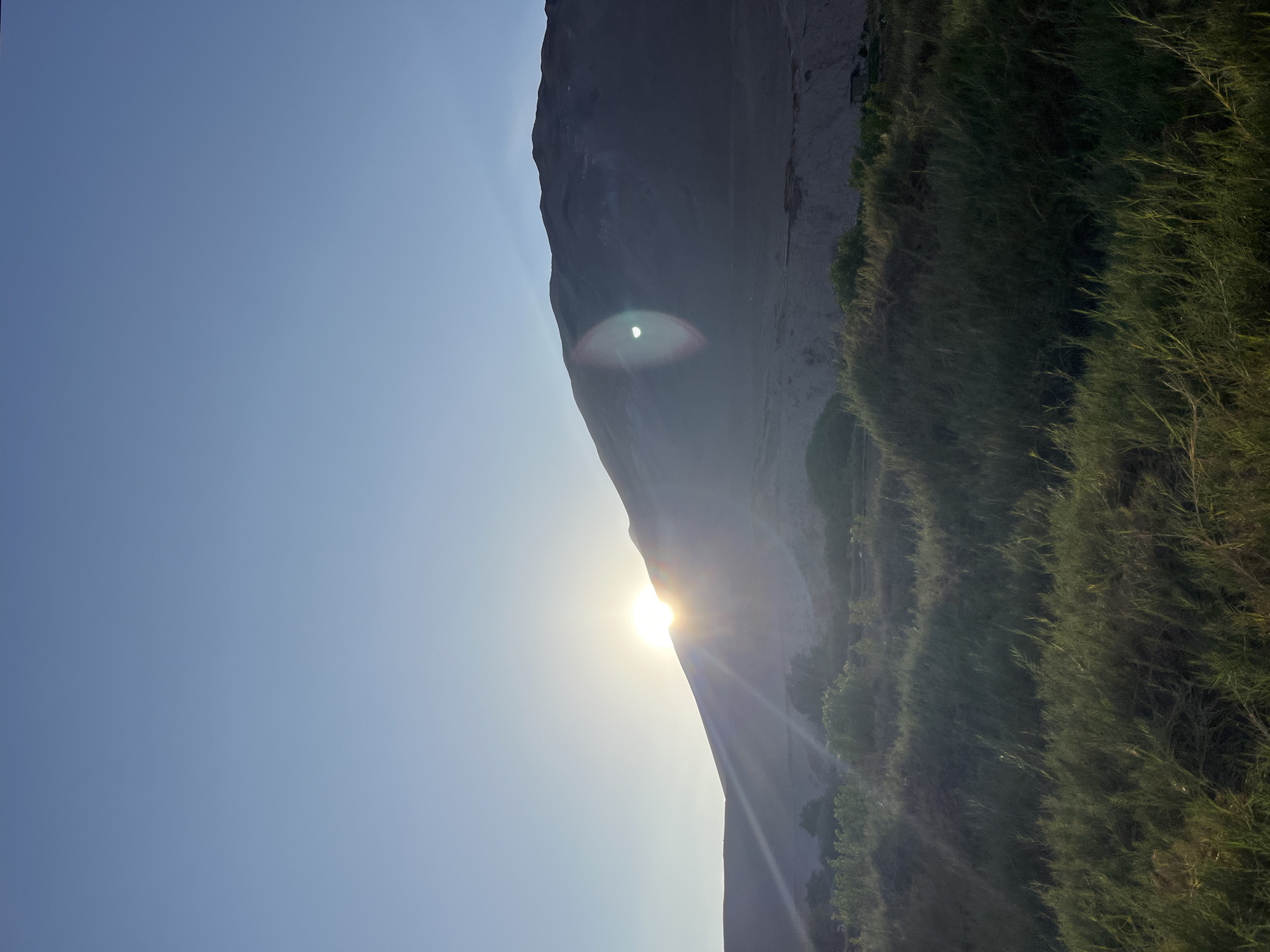 The image depicts the sun rising over El Morro de Arica, a steep hill in Arica, Chile. The sun's glare is visible between the edge of the hill and the sky. Vegetation is on the slope of the hill. The sky is a clear blue.