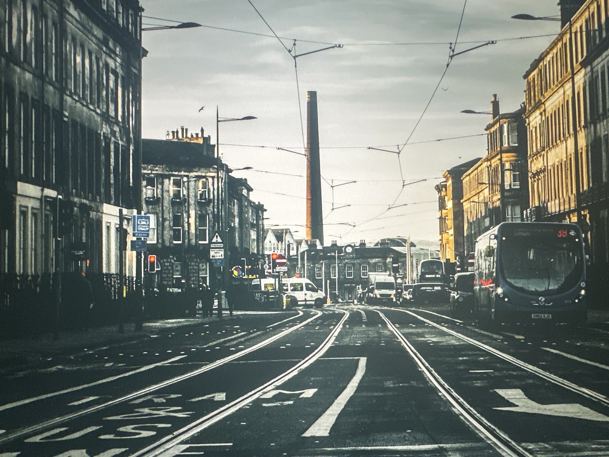 The image depicts a street scene with tram tracks in the foreground. On the left, a dark-colored building with many windows lines the street. On the right, a light-colored building with ornate architectural details stands. Several tram wires are visible overhead, supported by poles. In the middle distance, a tall brick chimney is prominent. Various vehicles, including a blue bus with the number "38" displayed, a white van, and other cars, are present on the street. The sky is overcast. The presence of tram tracks and the architectural style suggest an urban European setting, but the image does not provide specific details to identify the location as Bryansk, Russia.