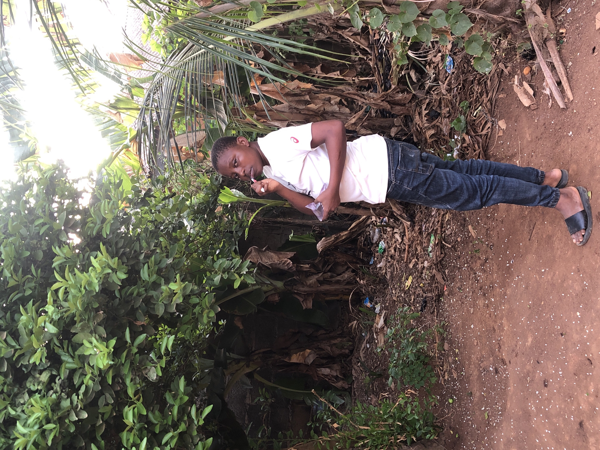 A young male is standing in front of a patch of dense foliage and dried foliage, holding a lollipop or similar treat to his mouth. He is wearing a white t-shirt with a visible red logo on the left shoulder, dark blue jeans, and black slides. The ground he stands on is dirt, with sparse vegetation growth and some scattered debris. The scene appears to be outdoors, possibly in a backyard or an unkempt roadside area, in the city of Unknown, Unknown.