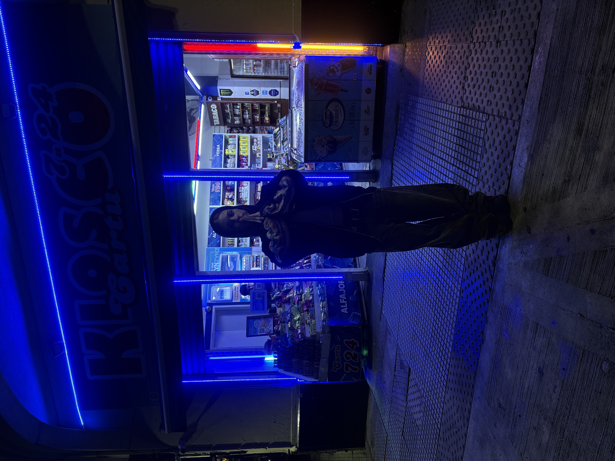 A person stands in front of a small kiosk with blue neon lighting, located on a patterned sidewalk. The kiosk, "Kiosco Cartu 7a24," sells various items including snacks, cigarettes ("TABACO"), and ice cream. A sign reads "ARCOR Helados CREMOSO." The person is standing near an ice cream cooler. The location is Buenos Aires, Argentina, as indicated by the Argentinian brands and kiosk style.