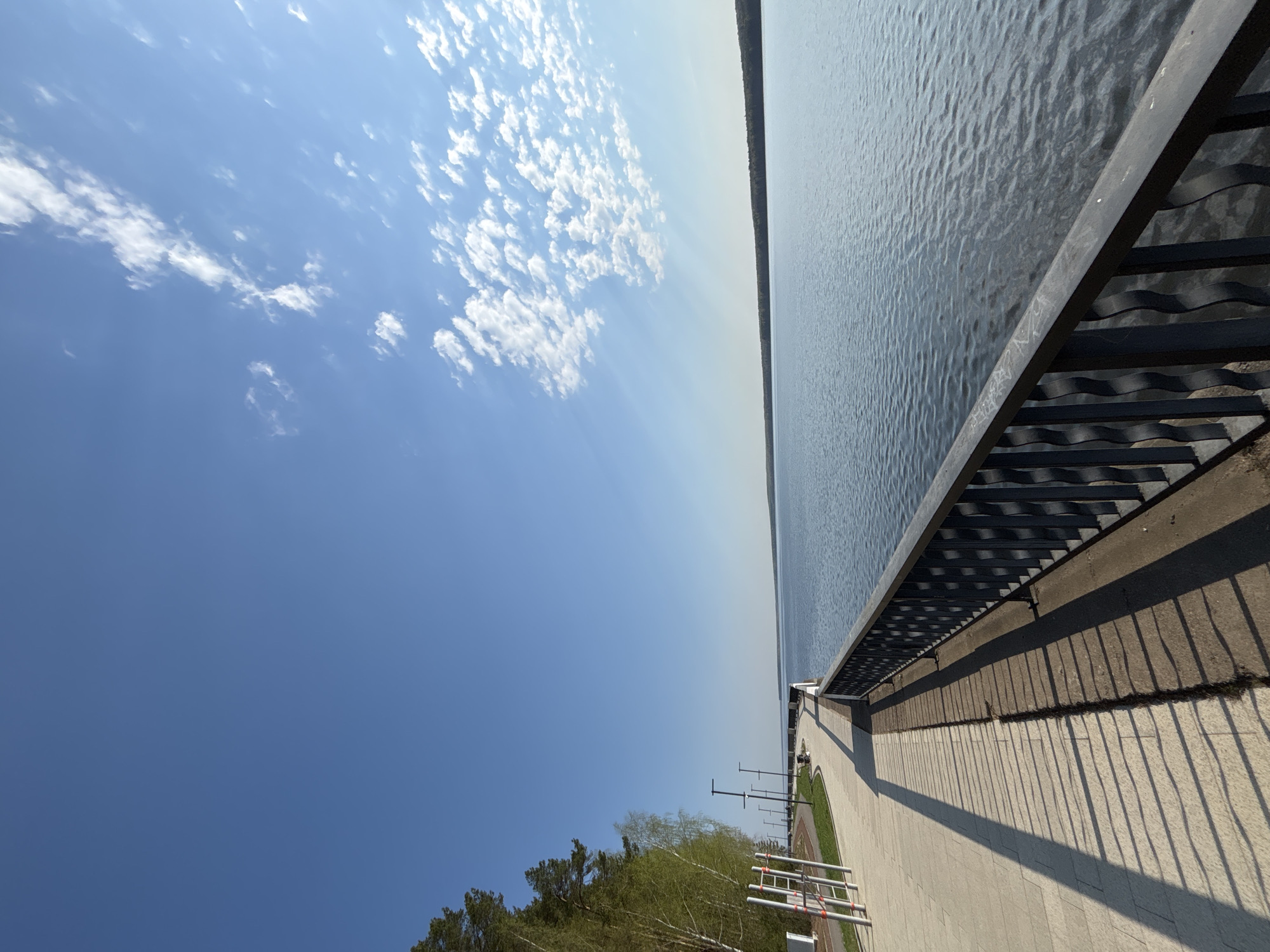 This image depicts a view from a promenade in the city of Chaikovsky, Russia, overlooking the Kama River. The sky is predominantly blue with scattered white clouds. To the right, a metal railing lines the edge of the concrete walkway, casting shadows onto the pavement. Beyond the railing, the surface of the Kama River is visible. In the lower portion of the image, trees are visible on the shoreline.