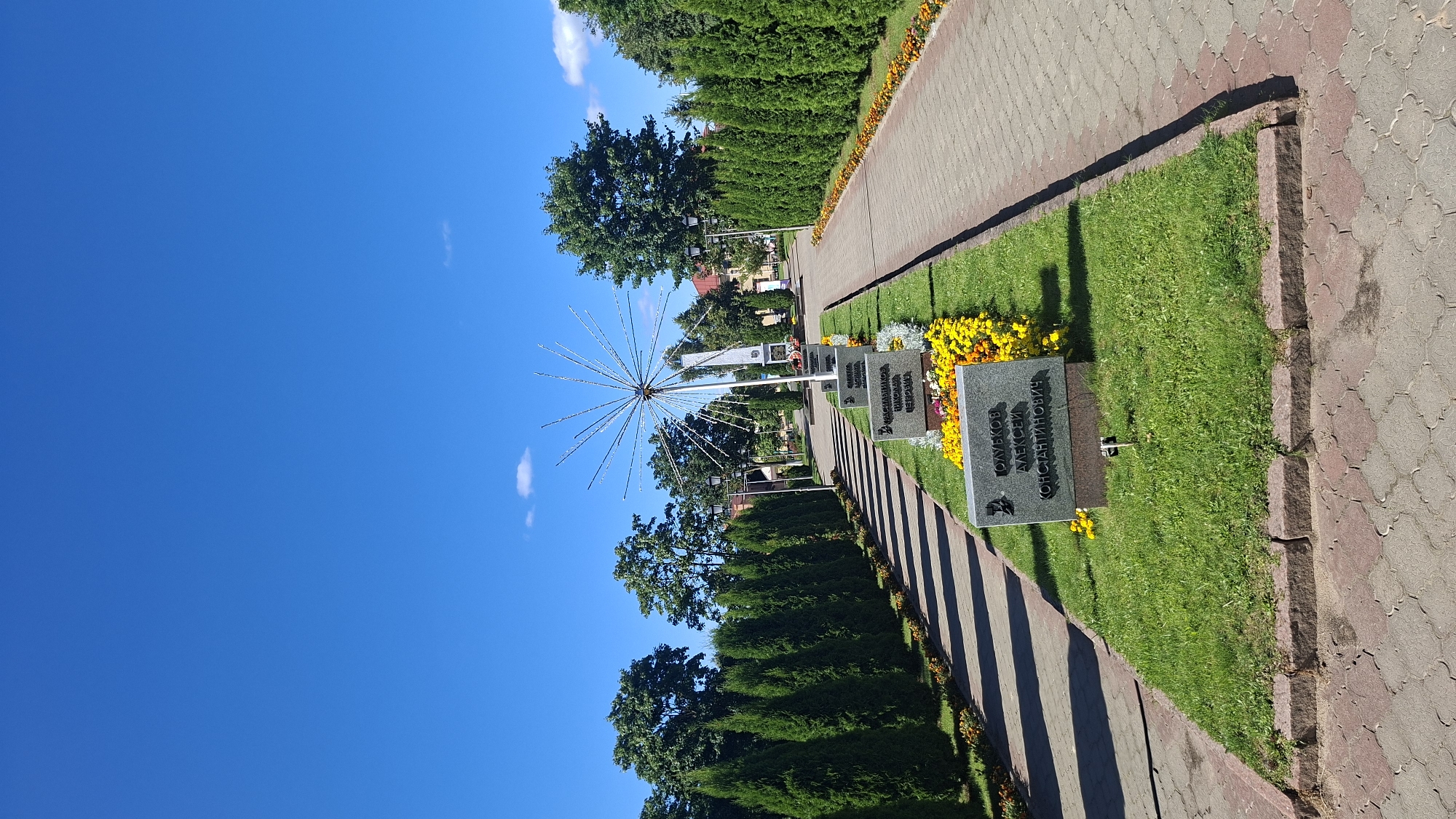 The image shows a memorial complex with several gravestones and flowering plants. A pathway made of paving stones runs through the scene, lined by rows of cypress trees. In the background, a sculpture with radiating elements stands tall, and behind it, a monument is visible. The sky is clear and blue with a few small clouds. The location is identified as the memorial complex in the city of Pastavy, Belarus, which honors fallen soldiers.