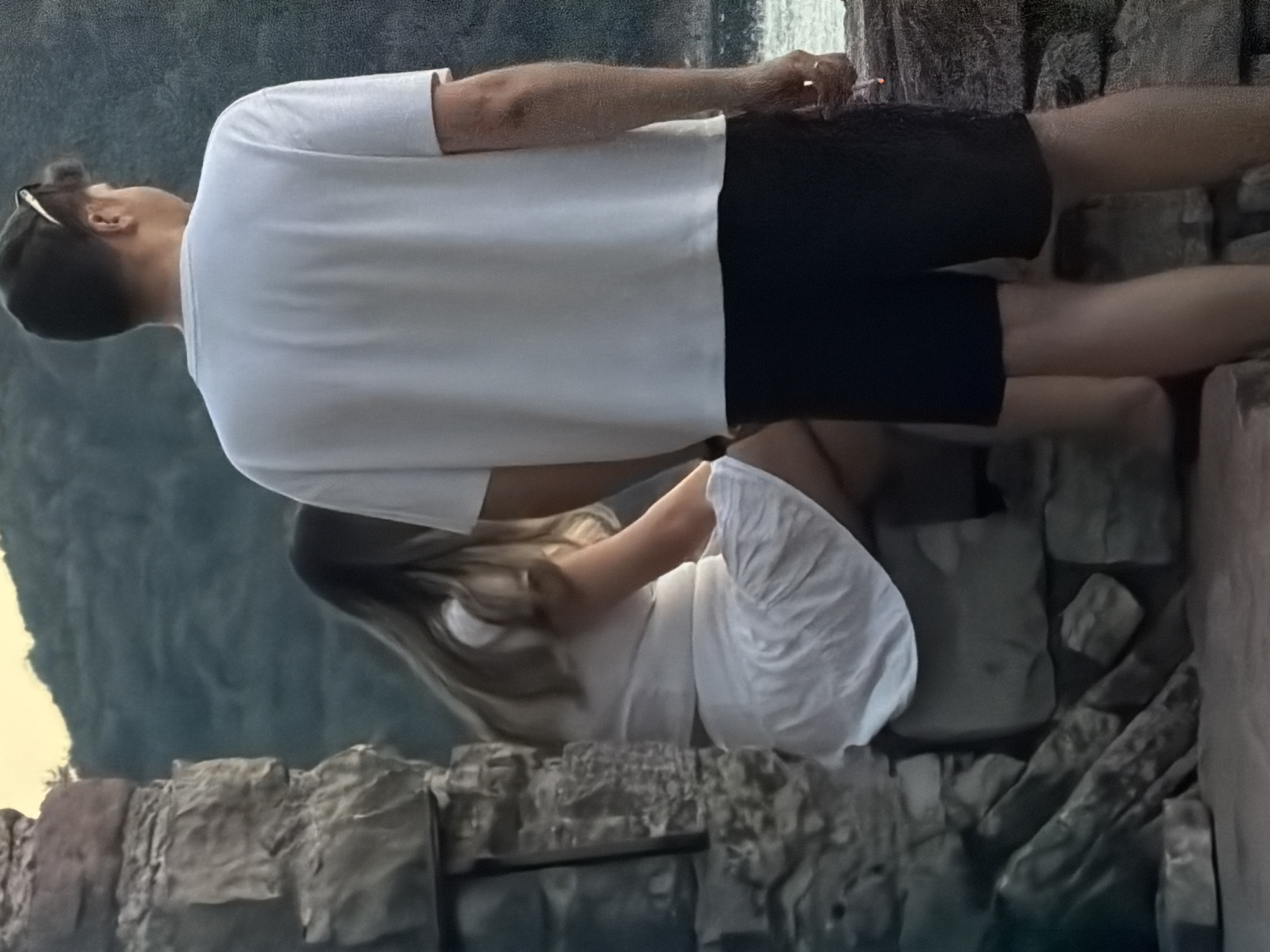 A young couple is posed outdoors, with a man in the foreground and a woman in the background. The man, viewed from behind, wears a white t-shirt and black shorts, with sunglasses pushed up on his head. He stands facing away from the camera, looking towards a body of water. The woman, also viewed from behind and to the left, is seated on a stone ledge. She wears a white dress and has long, light brown hair. The background shows a hilly, vegetated landscape and a body of water, likely the Adriatic Sea. The scene appears to be located near a stone structure, possibly part of an ancient ruin or fortification. The lighting suggests it is late afternoon or early evening. Given the context of Budva, Montenegro, this could be a scenic viewpoint overlooking the coastline or a historic site within the city.