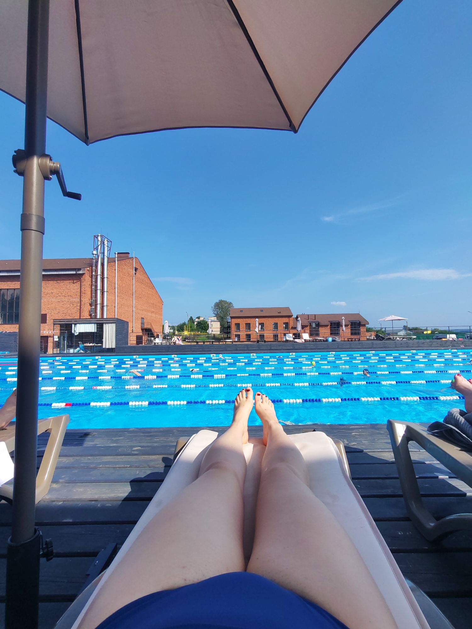 The image depicts a sunny day at an outdoor swimming pool facility, likely located in Pirogovo, Russia. In the foreground, a person is lying on a lounge chair, with their legs extended and bare feet visible. The person is wearing a blue garment, possibly a swimsuit. To the left, an umbrella provides shade, with its pole and crank mechanism partially visible. The lounge chair is situated on a wooden deck.
Beyond the deck is a bright blue swimming pool with lane markers. Several people are visible in the pool, some swimming or engaging in water activities. In the background, brick buildings with large windows are present, possibly part of the pool facility or related structures. The sky is a clear, vibrant blue with a few wispy clouds. The overall atmosphere suggests a recreational setting on a warm day.