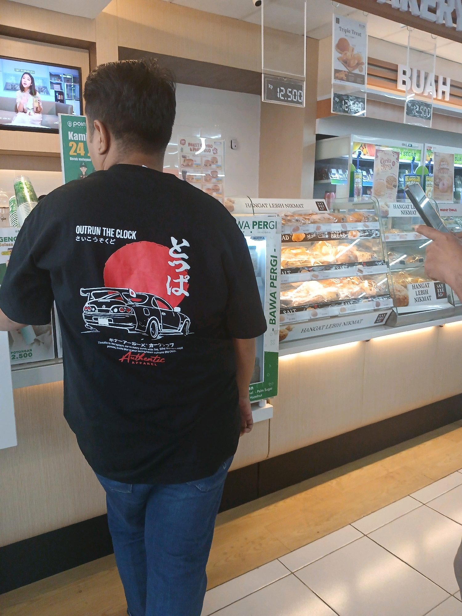 In Depok, Indonesia, a person wearing a black t-shirt with a graphic of a red sun and a white car is standing in front of a display of baked goods. The t-shirt has Japanese characters and English text that reads "OUTRUN THE CLOCK" and "Throttle to the horizon, curve gently curve with fire. Who needs more, peace, limits, blur your racing heart outruns the clock. Authentic Apparel". The display case contains various pastries, some individually wrapped. Above the display, there are signs with prices and promotional text in Indonesian, such as "HANGAT LEBIH NIKMAT" (Warm is more enjoyable) and "BAWA PERGI" (Take away). In the background, a television screen shows a woman with a drink. The overall setting appears to be a food establishment or convenience store.