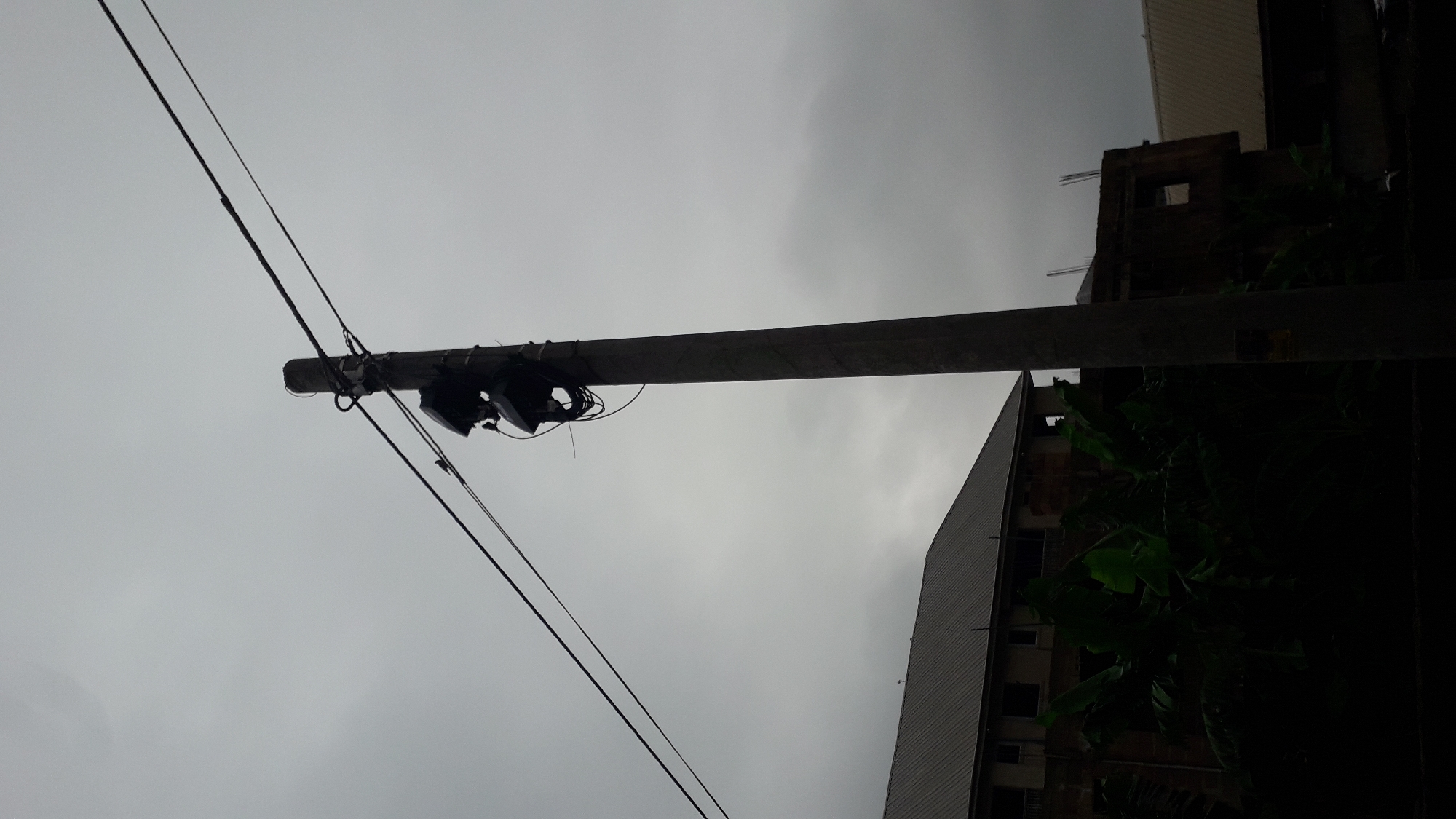 The image shows a low-angle view of a utility pole in Nkpor, Nigeria. Two power lines are strung across the pole. Attached to the pole are what appear to be two black security cameras. In the background, a building with a metal roof is partially visible, along with a building under construction with plants growing on its facade. The sky is overcast, suggesting poor weather conditions.