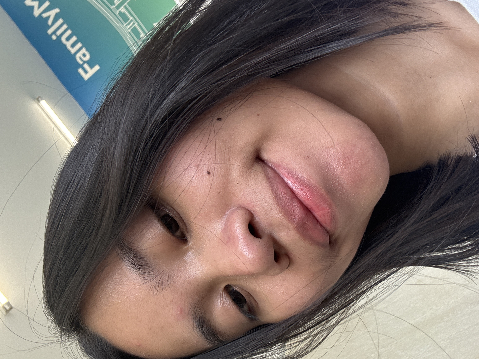 The image is a close-up selfie of a young woman with dark hair and medium-toned skin. She is looking down and slightly to the left. Her lips are slightly parted, and she appears to be smiling faintly. There is a small red blemish on her forehead, and a few moles are visible on her cheek. Her hair is dark and appears to be straight, falling around her face and shoulders.
In the background, a blue and green sign with white lettering that reads "FamilyM" is partially visible. This signage is consistent with branding found in commercial establishments. The lighting suggests an indoor environment, possibly a store or mall. The overall context and the presence of the "FamilyM" sign indicate a location within East Jakarta, Indonesia, likely a retail area or a shopping center.