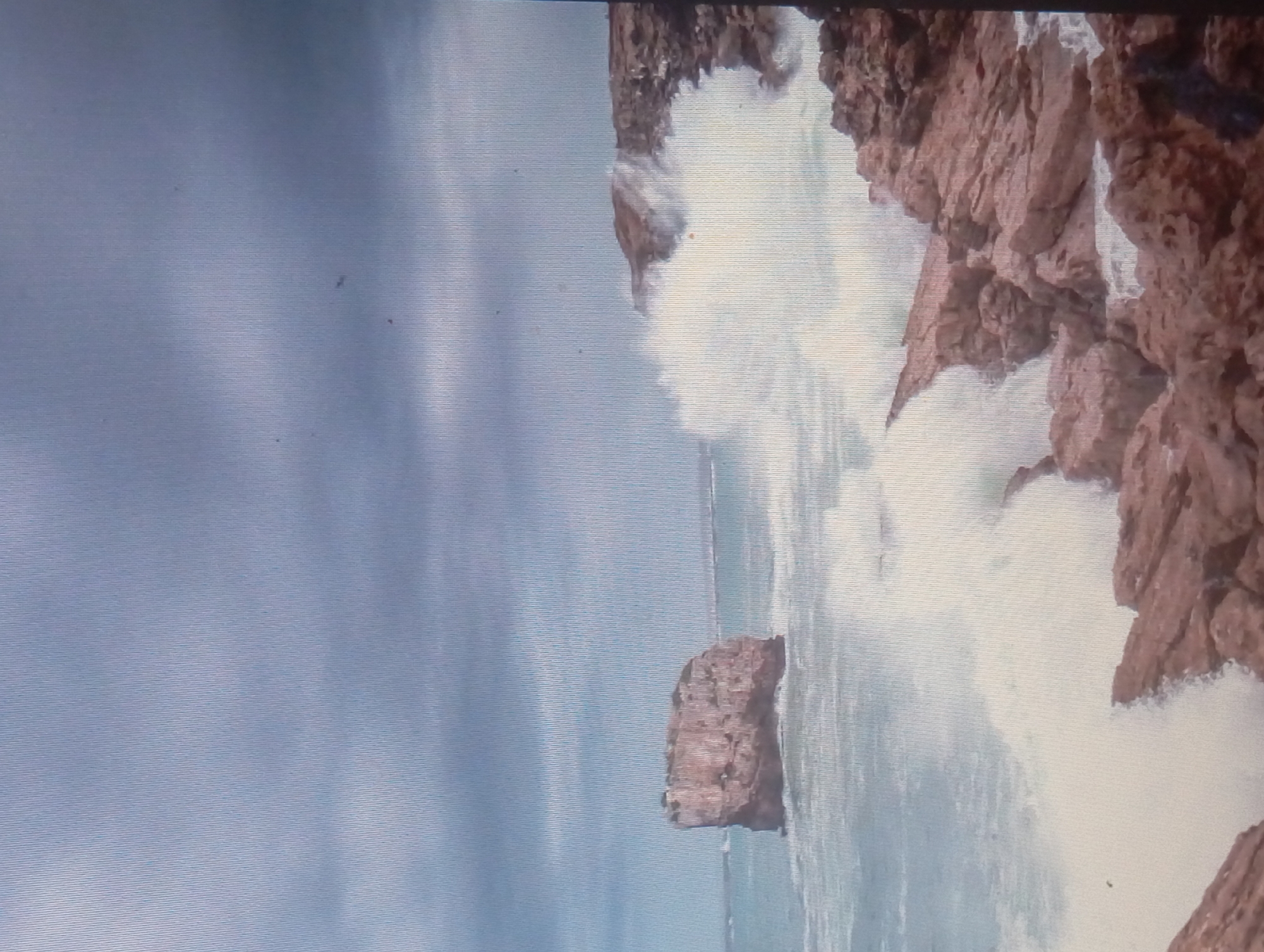 The image depicts a coastal scene with rocky cliffs and ocean waves. White surf crashes against the reddish-brown rocks, creating a dramatic spray. In the background, there is a large rock formation in the sea, possibly an island or large boulder. The sky is overcast with grey clouds. The image does not appear to be in Karaganda, Kazakhstan, as it is a landlocked region with no coastline. The location is more likely to be somewhere along a sea coast.