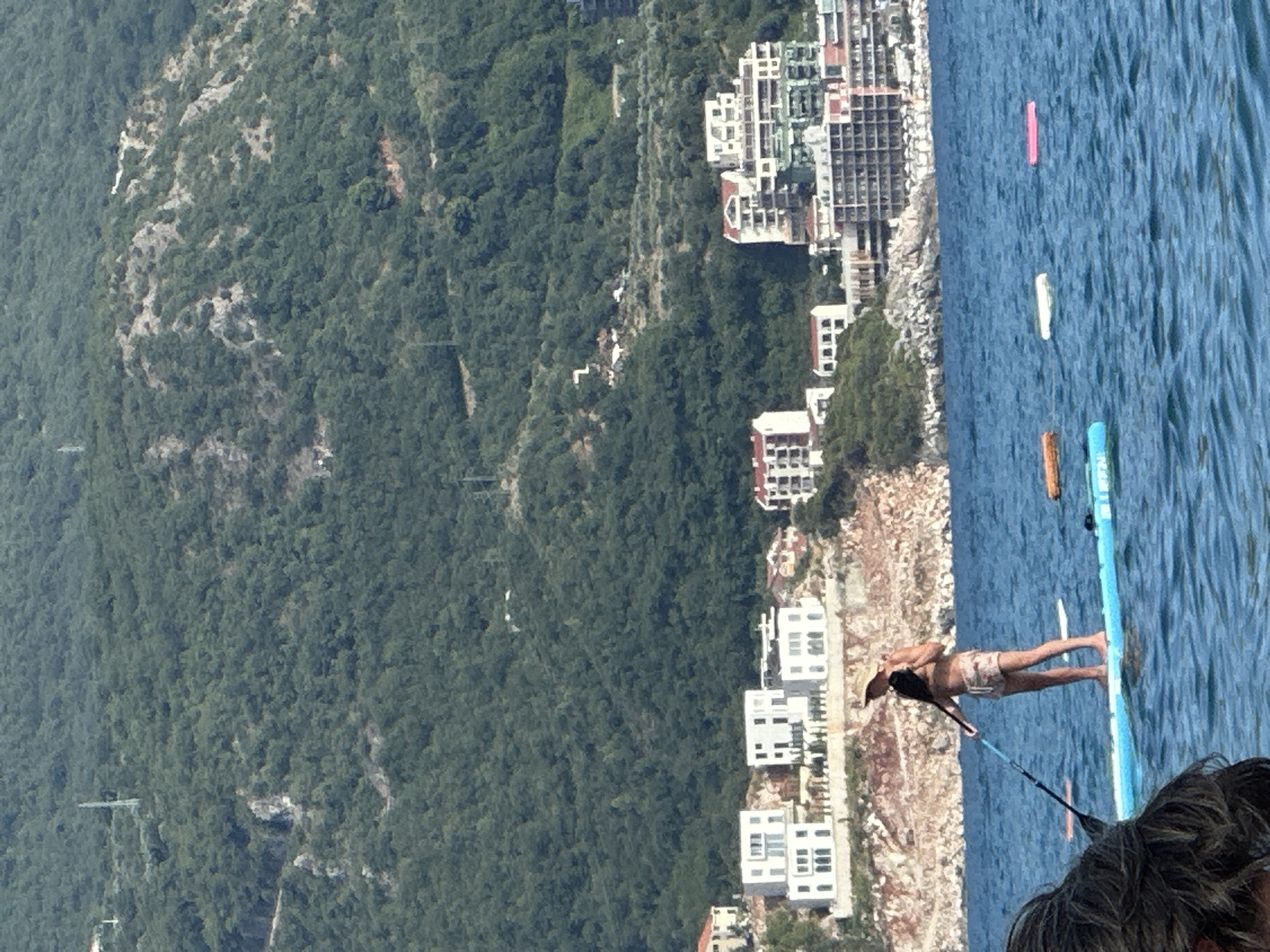 A person is paddleboarding on the blue sea, facing away from the camera. The person is wearing a straw hat and patterned shorts, holding a paddle. The paddleboard is light blue. In the background, there are buildings on a rocky incline leading up to a densely forested mountain. The buildings are modern, with white facades and multiple stories. The sea is calm with some ripples. There are other inflatables in the water. The location is Boreti, Montenegro.