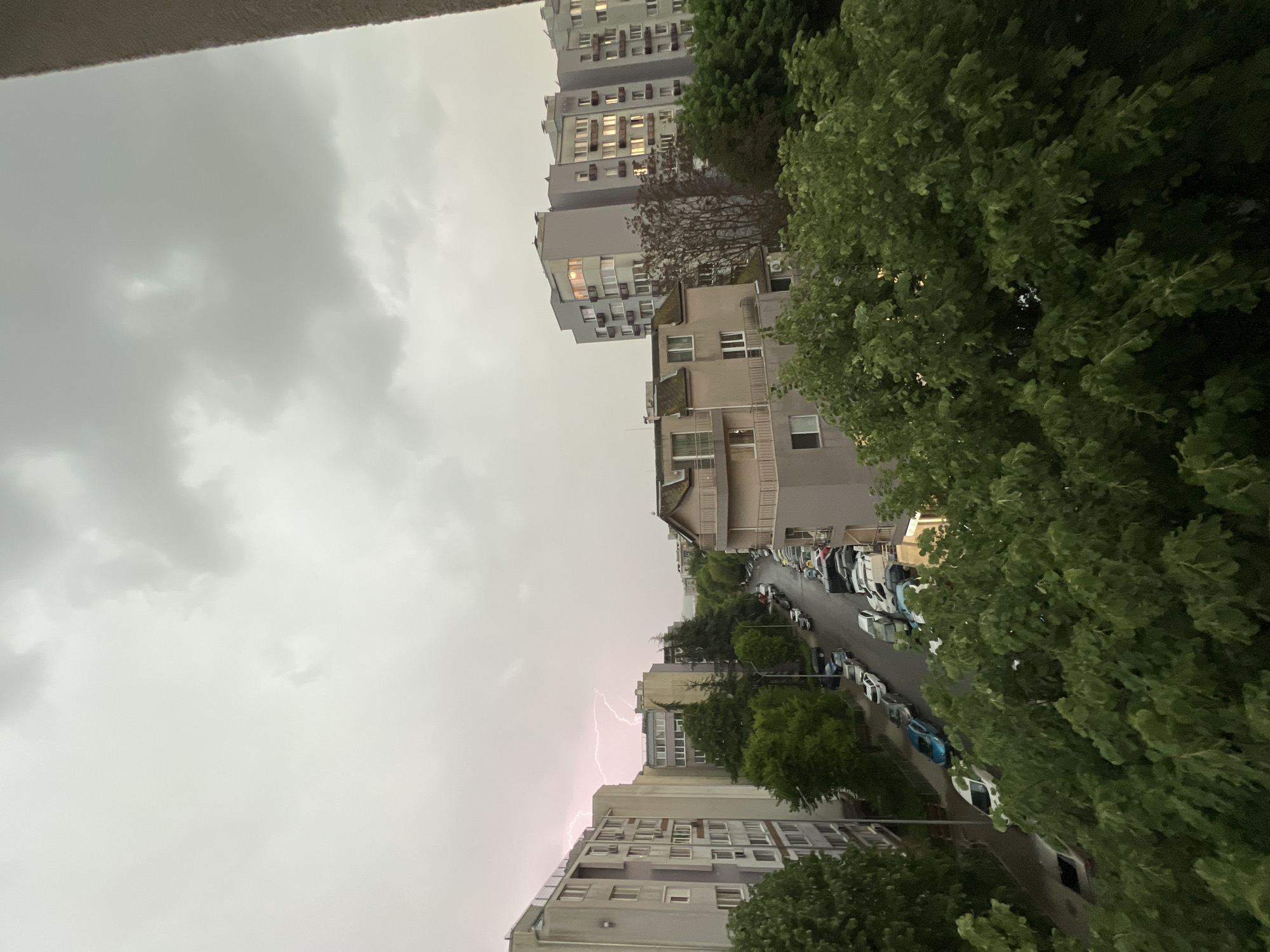 The image depicts an urban scene during a thunderstorm in Burgas, Bulgaria. Gray apartment buildings line a street filled with parked cars. Dense green trees partially obscure the view. Lightning flashes in the sky between the buildings. The sky is overcast with gray clouds.