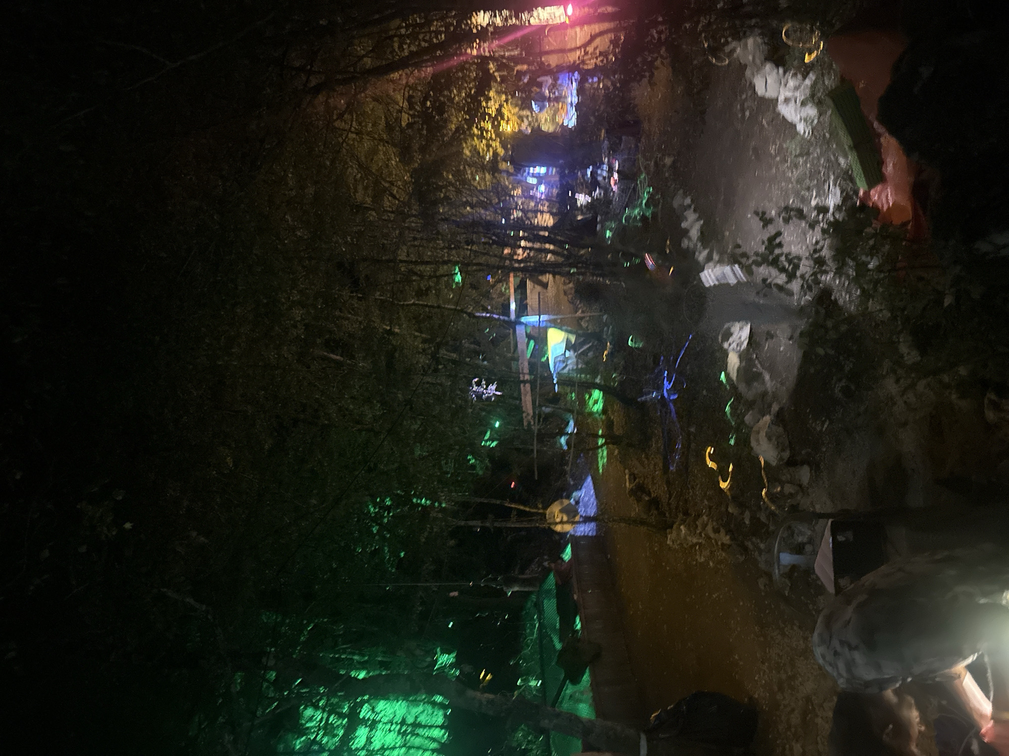 The image depicts an outdoor scene at night, likely in a wooded area with some artificial lighting. The environment is characterized by trees and foliage, with pathways or cleared areas visible. Various colored lights, including green, blue, yellow, and pink, are illuminating sections of the scene, suggesting some form of decoration or event.
In the foreground, the lower left of the image shows a dark, blurry object that may be a person or a large item. Further into the scene, there are structures that appear to be made of wood or fabric, possibly temporary shelters or stages, illuminated by colored lights. There are also rocks and scattered debris visible on the ground.
The lighting creates a somewhat surreal and atmospheric effect, with some areas brightly lit and others in deep shadow. The overall impression is of an illuminated outdoor gathering or display at night. The location is identified as Bukovik, Montenegro, though no specific landmarks of the city are discernible in the image.