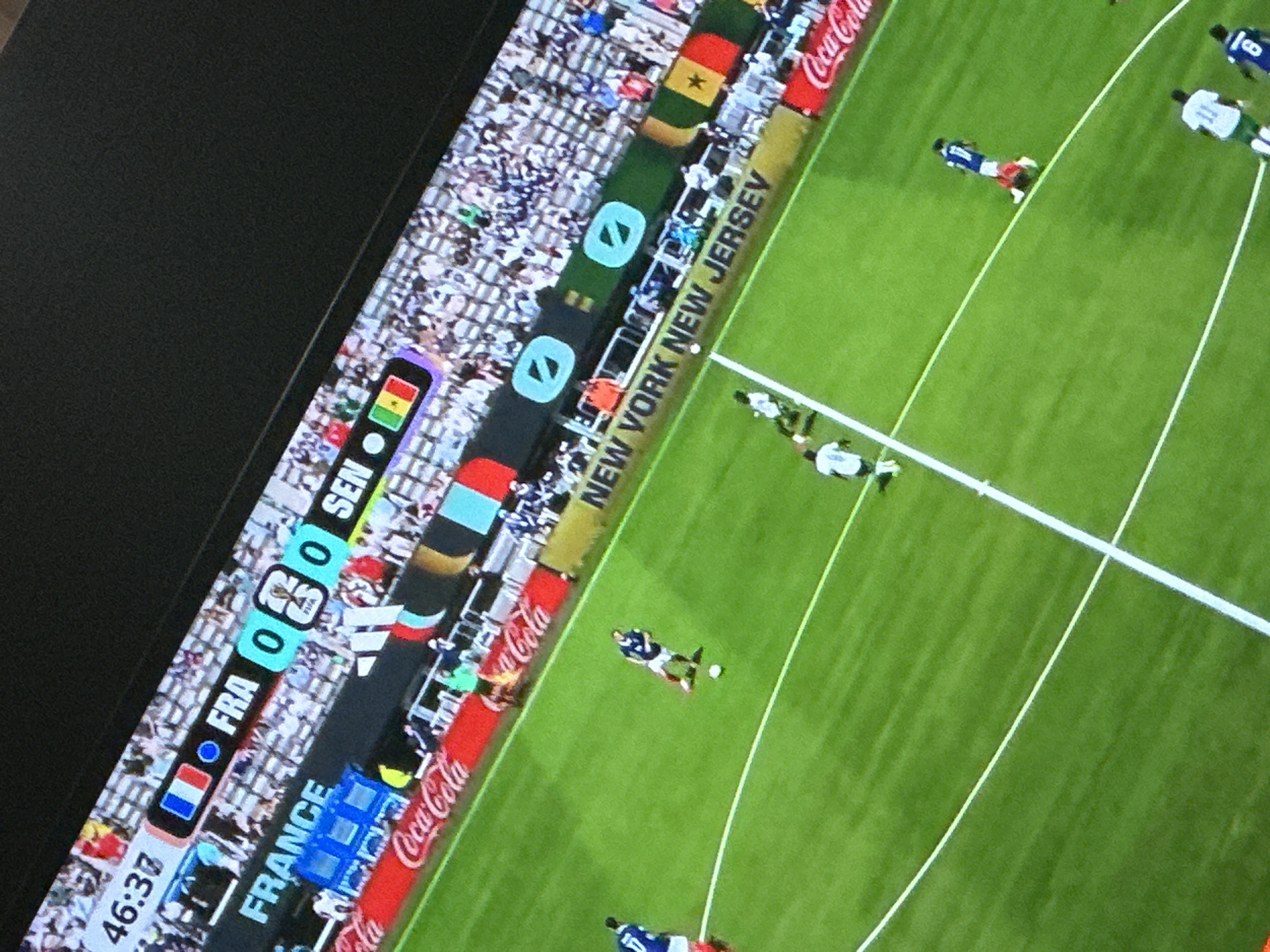 The image shows a soccer match being played on a green field with white boundary lines. There are spectators in the stands behind the field. A large screen at the top of the image displays game information, including the score, which is 0-0, and the teams, France (FRA) and Senegal (SEN). The time shown is 46:37. The advertisement boards around the field show "Coca-Cola" and "New York New Jersey". Several soccer players in white and blue uniforms are on the field, some actively playing and others in posed positions. The overall scene depicts a professional soccer game in progress. There is no indication of the city of Rayfield, Nigeria.
