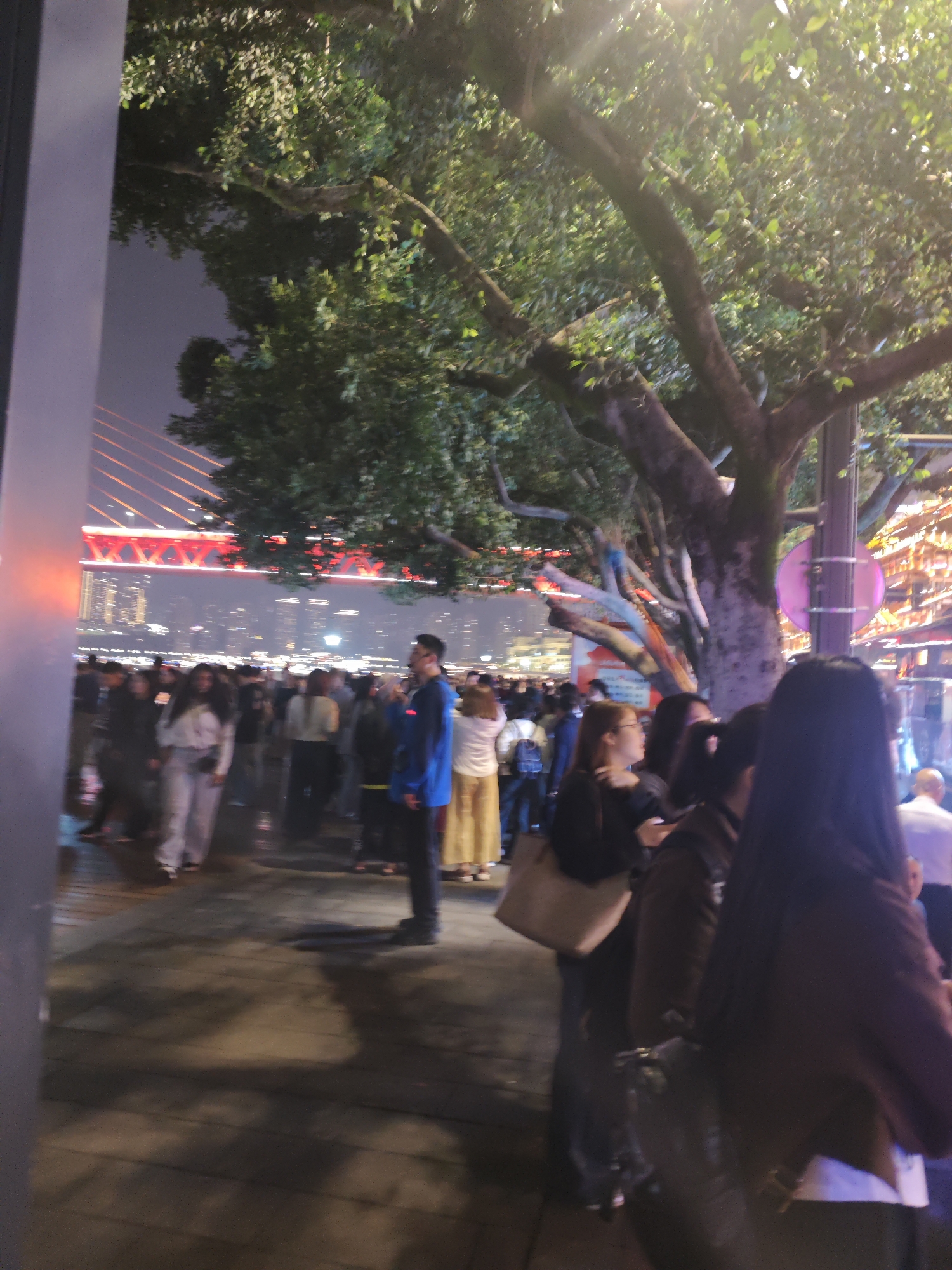 The image depicts a crowded outdoor scene at night in Yuzhong District, China. A large group of people is gathered on a pedestrian walkway, with a bridge illuminated in red in the background. The walkway is lined with trees and streetlights. A building is visible in the distance behind the bridge.