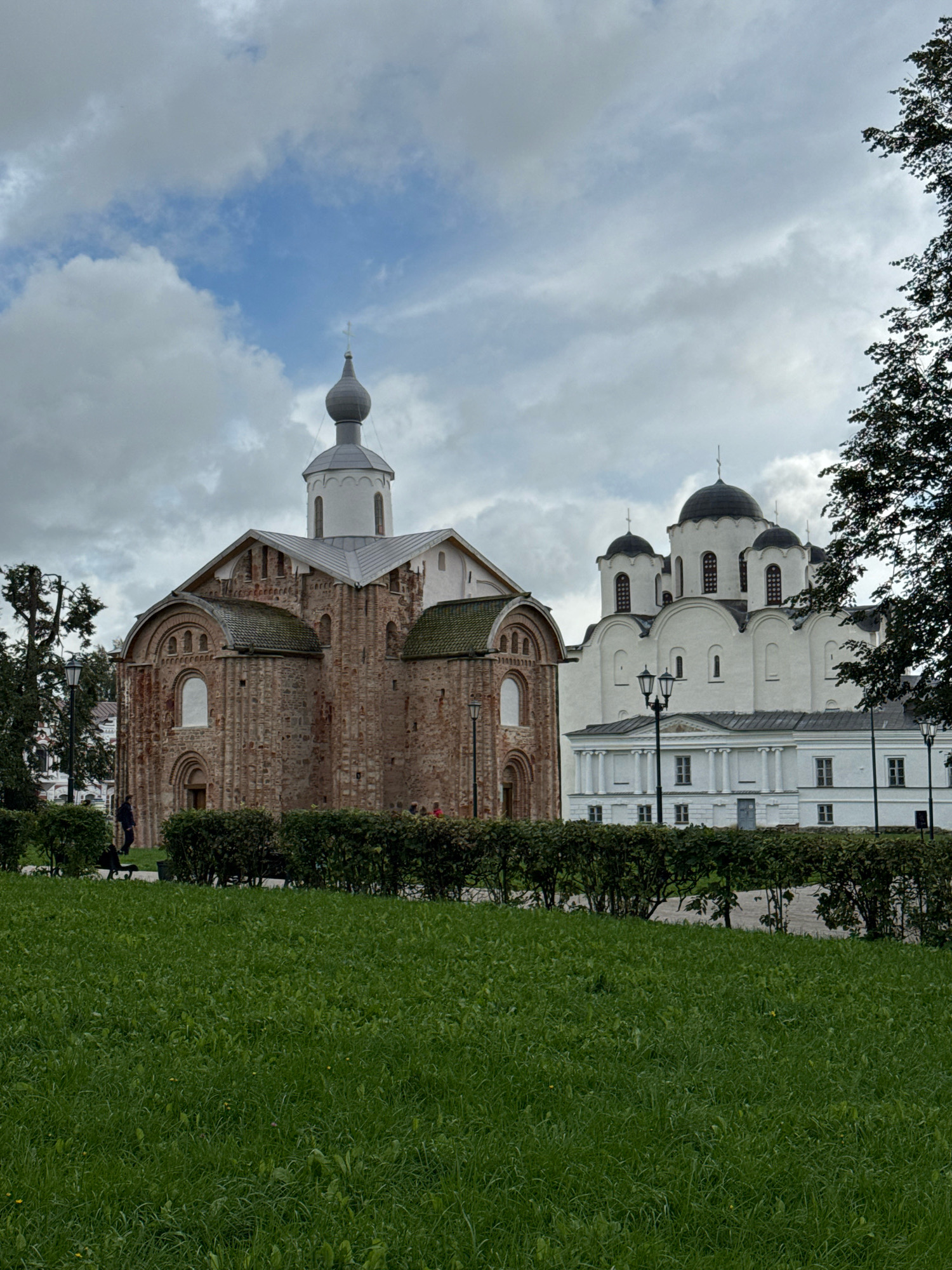 The image shows the Saint Sophia Cathedral and the Bell Tower of Saint Sophia Cathedral in Veliky Novgorod, Russia. The Saint Sophia Cathedral is a large, white church with multiple domes. The Bell Tower is a tall, slender structure with a grey dome. In front of the cathedral and bell tower is a grassy lawn with trimmed bushes. The sky is cloudy. There are a few people walking around the grounds.