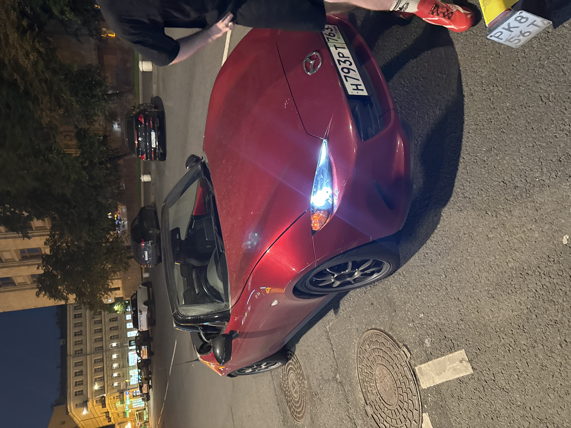 The image shows a red Mazda convertible parked on a city street at night. The car has its headlights on and is facing towards the viewer's right. The license plate reads "H793PT 763". A person wearing a black t-shirt and black shorts, and red sneakers, is standing next to the car, partially visible on the right side of the frame. Behind the car, other vehicles are parked along the street, and buildings can be seen in the background. The scene appears to be in an urban area, likely in Moscow, Russia, given the license plate format and Cyrillic script on a nearby license plate fragment at the bottom right. There are manhole covers on the asphalt road. The lighting suggests it is evening or night, with streetlights and lights from the buildings illuminating the scene.