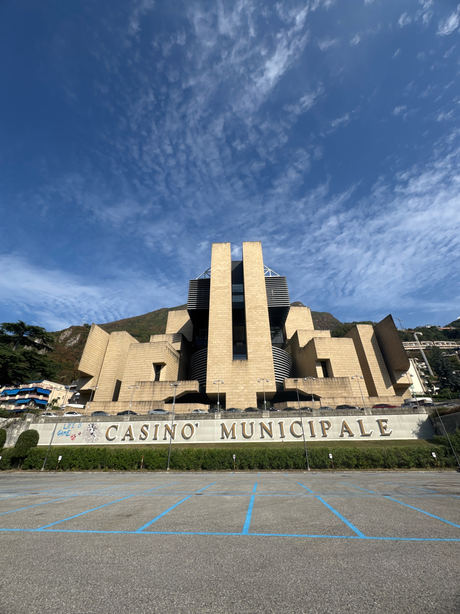 The image shows the Casino Municipale in Campione d'Italia, Italy, on a sunny day with a partly cloudy blue sky. The building's distinctive architecture features large, angular concrete structures with a beige facade. In front of the building, a row of cars is parked along a retaining wall. Below this wall is a large, empty asphalt parking lot marked with blue parking lines. A sign with the words "LIFE IS GAME" and "CASINO' MUNICIPALE" is visible above a neatly trimmed hedge. Green trees and a hillside with buildings are visible in the background.