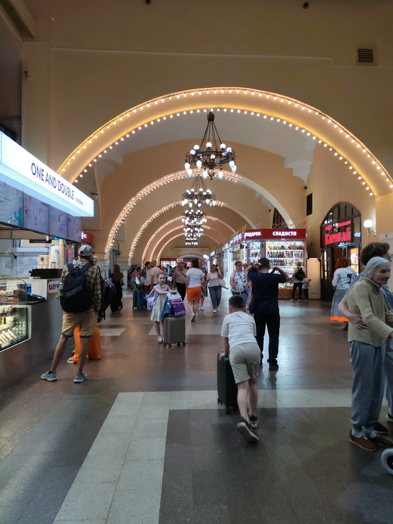 The image depicts a bustling interior walkway, likely a train station, in Moscow, Russia. Arched ceilings, adorned with strings of fairy lights and ornate chandeliers, create a grand architectural style. The walkway is populated with numerous individuals, many of whom are carrying luggage, suggesting travel. Shops line the sides of the walkway, with visible signage for "ПОДАРКИ" (Gifts) and "СЛАДОСТИ" (Sweets), indicating retail establishments. One shop prominently displays the Cyrillic word "КАРАВАЙ" in red letters. The floor is a polished stone or tile surface, reflecting the ambient light. People are shown walking, some in groups, while others are alone, engaged in various activities such as looking at their phones or pushing suitcases. The overall atmosphere is one of movement and commerce within a public transit hub.