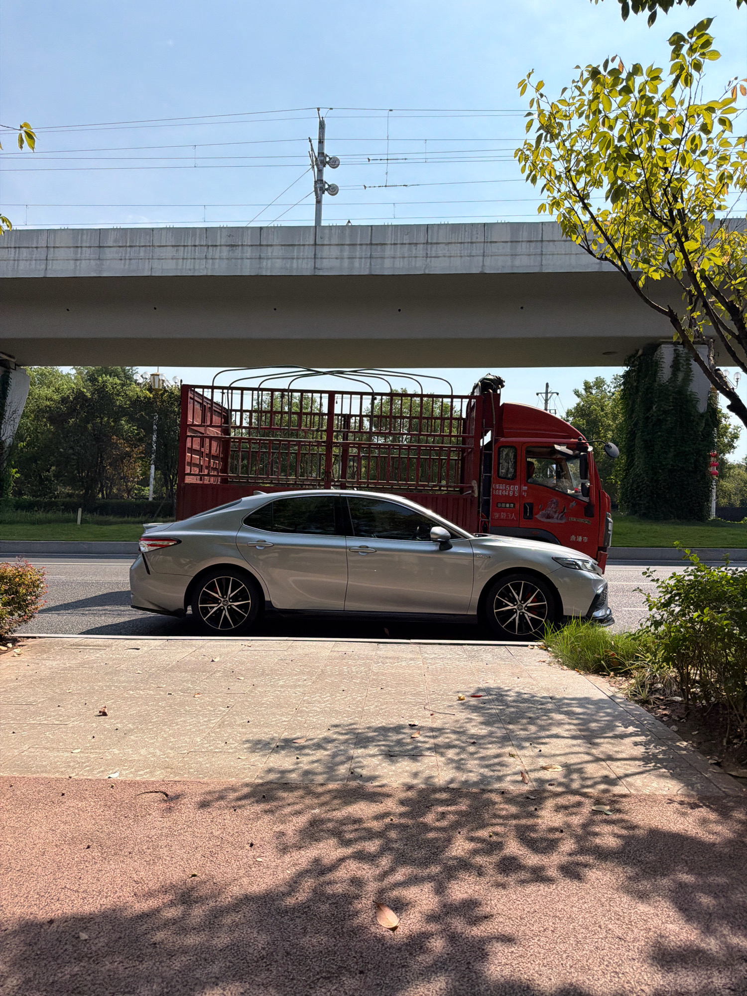 A silver sedan is parked on the side of a road, adjacent to a sidewalk, under an elevated structure. Behind the sedan is a red truck with a caged bed. The truck has Chinese characters on its door. The elevated structure appears to be part of a bridge or overpass. There are trees and greenery visible in the background, as well as power lines and a utility pole. The sky is clear and blue. The scene is lit by sunlight, casting shadows on the sidewalk. The context suggests this image was taken in Jiangjin District, Chongqing, China, a location known for its infrastructure and surrounding natural landscape.