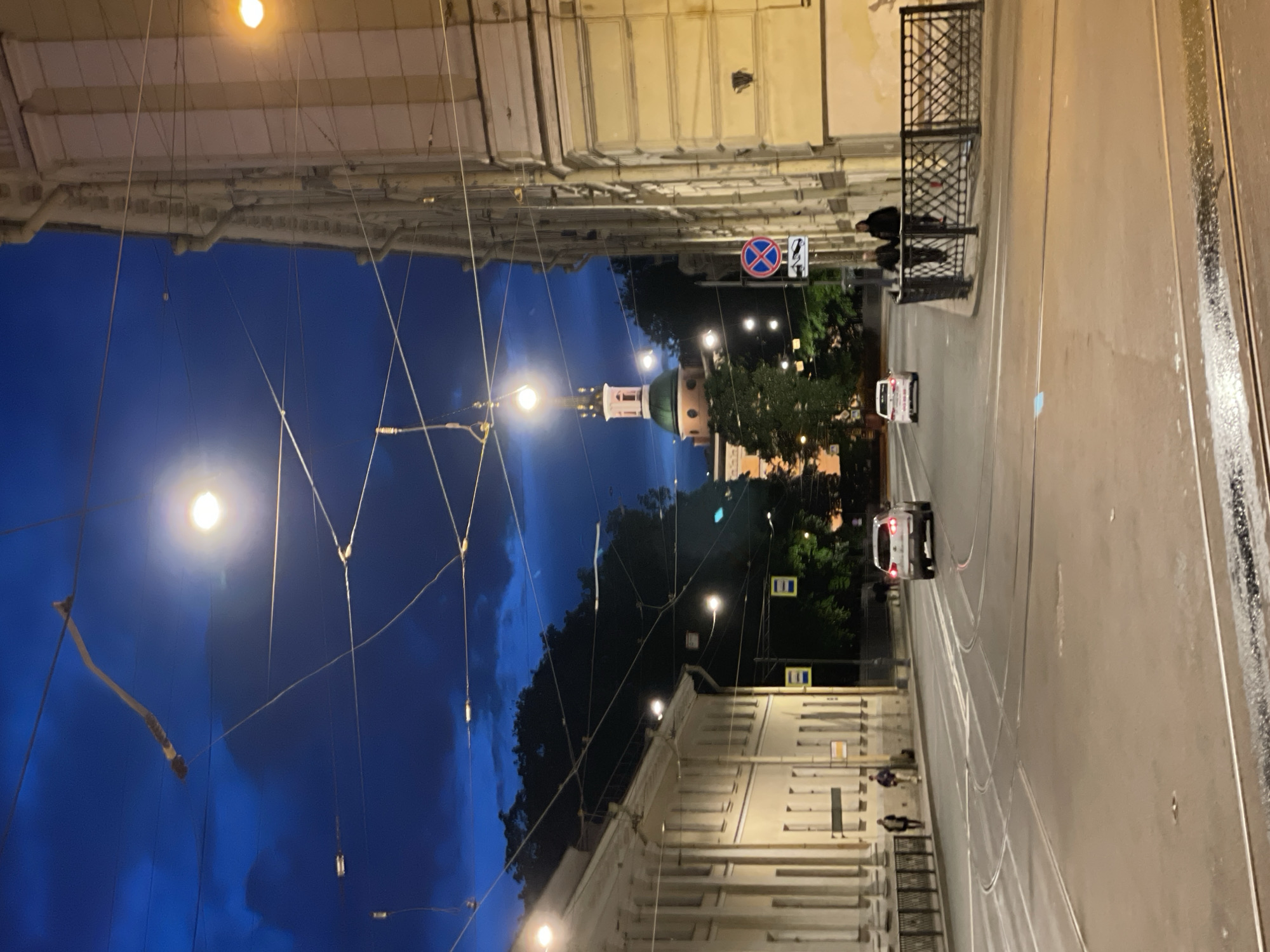 The image captures a nighttime or late twilight street scene in Saint Petersburg, Russia. The perspective is looking down a city street, flanked by tall, classical-style buildings on both sides that are softly lit by artificial light. Their ornate facades and multiple windows are visible.
In the distance, a prominent, round architectural tower with a domed, green roof and a spire rises above the surrounding trees and buildings, acting as a focal point. Numerous overhead wires, likely for trams or trolleybuses, crisscross the dark blue sky, which shows hints of darker clouds. Streetlights illuminate these wires, creating glowing points of light.
The road surface appears wet, possibly from recent rain, reflecting the streetlights. Tram tracks run along the length of the street. Two white cars are visible on the road; one in the middle distance appears to be a taxi with a light on its roof.
On the sidewalks, several people are present. One individual is seated on a bench or barrier near a black metal fence on the right side of the street. Other figures are seen walking further down the street on the left.
Notable details include a red and blue "no stopping" sign (red cross over blue circle) and a blue and white "no parking" sign (blue circle with a red diagonal line through a white 'P') on the right side of the road. Yellow rectangular signs with black symbols are also visible but unreadable. The overall mood is quiet and atmospheric, characteristic of an urban evening.