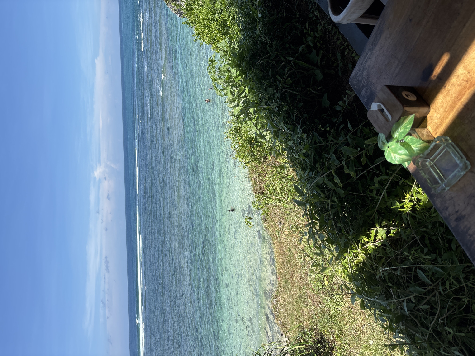 The image depicts a coastal scene, likely in Kuta Selatan, Indonesia. The clear, turquoise water of the ocean is visible, meeting a shoreline densely covered with green vegetation and grass. Two figures are visible in the water. On the right side of the frame, a wooden table is partially visible, with a small square glass container containing a plant. Next to the plant is a wooden box. The sky is blue with some clouds. The image is oriented vertically.