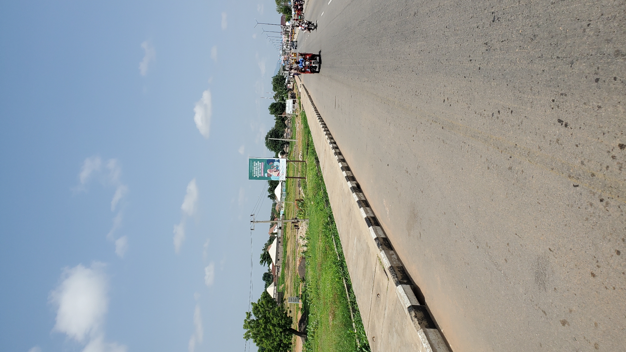 The image depicts a street scene in Jalingo, Nigeria, on a bright, sunny day with scattered white clouds in a light blue sky. The foreground is dominated by a grey asphalt road with some oil stains and faint tire tracks. To the left, a concrete sidewalk with a black and white striped curb runs along the edge of the road. Beyond the sidewalk, a strip of green grass and vegetation is visible, with a large tree on the left side. In the background, a billboard stands on the right side of the road, displaying text and an image. Further down the road, several buildings with conical roofs are situated in a semi-rural setting. Traffic is present on the road, including a three-wheeled vehicle (a Keke Napep) with passengers and a motorcycle. Streetlights are visible lining the road in the distance. The scene appears to be a typical urban or peri-urban environment in Nigeria, characterized by the presence of local transportation and common architectural styles.