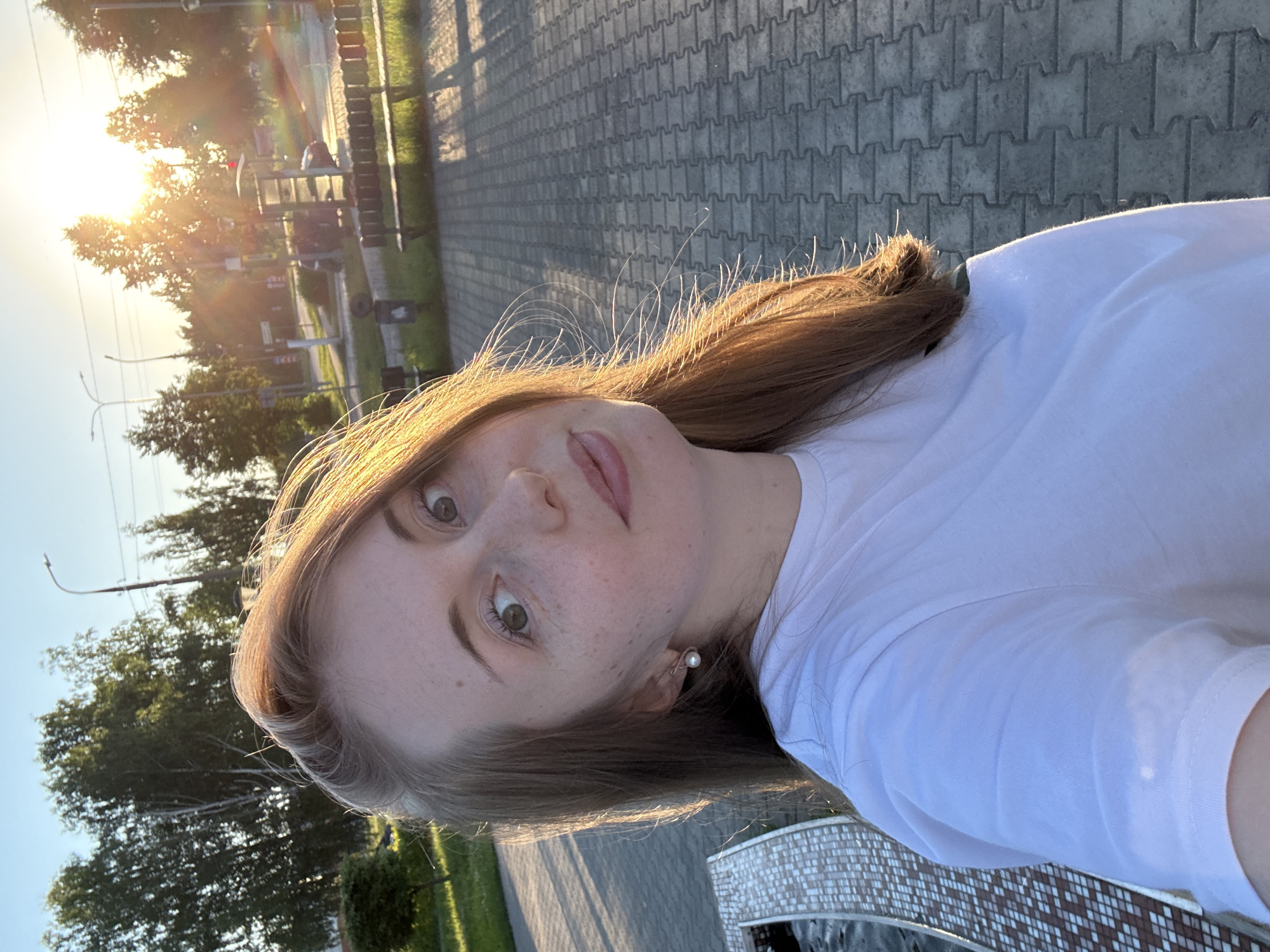 A young woman with long, auburn hair and light skin is taking a selfie outdoors. She is wearing a white t-shirt and a pearl earring. The background shows trees, a street with a bus stop, and a paved walkway. The sun is setting, casting a warm glow on the scene. The woman's face is turned towards the camera, and she has a slight smile. The image is a medium shot, with the woman's head and shoulders filling most of the frame. The location appears to be a park or public area in a city.