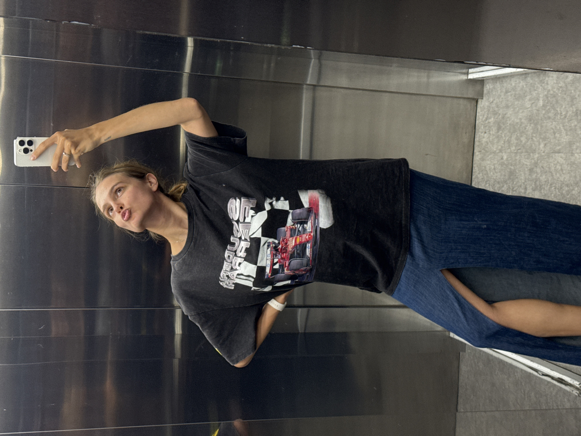 A blonde woman in blue jeans and a graphic tee is taking a selfie with her iPhone in an elevator. The location is Unknown, Unknown.