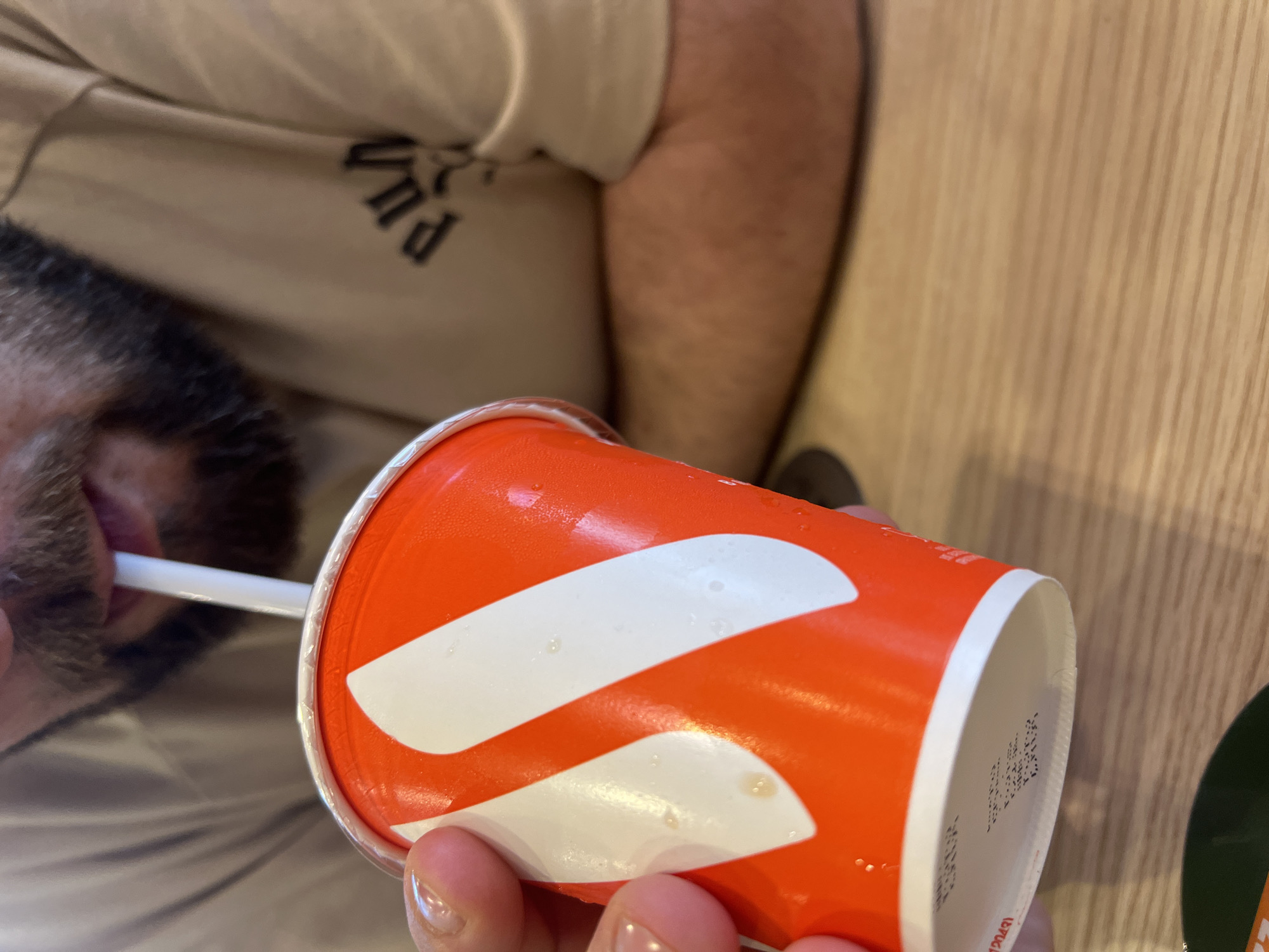 The image shows a close-up of a person drinking from an orange and white disposable cup with a white straw. The person's mouth and beard are visible, as is their hand holding the cup. The background shows a beige t-shirt with a black graphic design and a wooden surface, likely a table. The image appears to have been taken indoors, possibly in a restaurant or cafe. There are condensation droplets on the cup. The image does not contain any information or context that would allow for determining the location in Yekaterinburg, Russia.