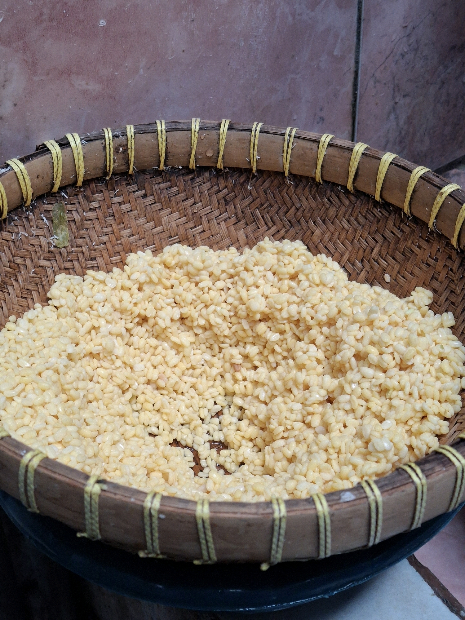 In Lumajang, Indonesia, a close-up, eye-level shot shows a woven bamboo basket filled with split mung beans. The basket has a thick rim reinforced with yellow twine. The mung beans are a pale yellow and are piled high in the basket, with some appearing to be slightly moist. The background consists of a textured, pinkish-brown wall.