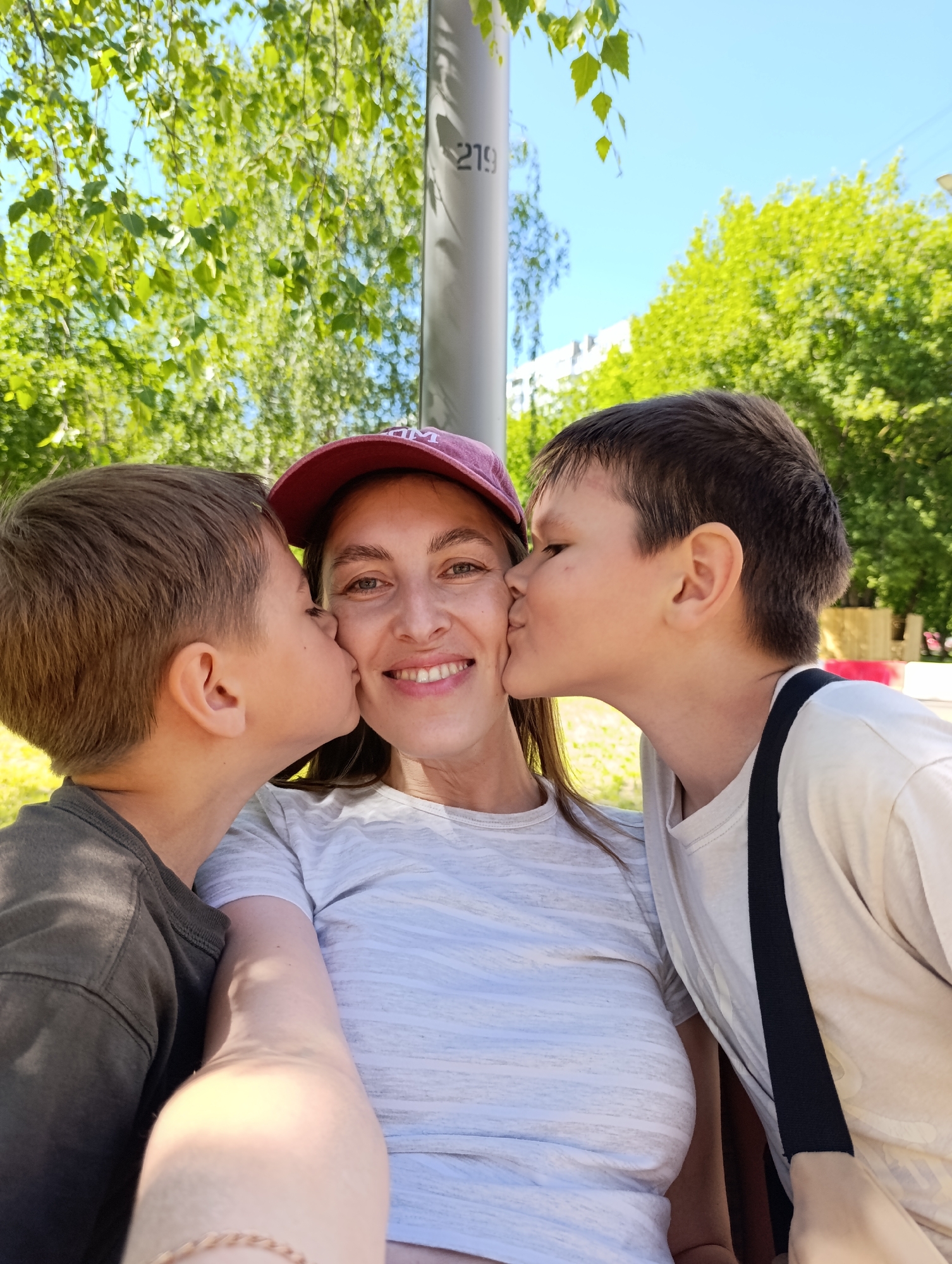 This high-resolution digital photograph captures a close-up, selfie-style shot of an adult female with two male children, likely her sons, in an urban green space. The adult female, positioned centrally, is smiling broadly, revealing her teeth. She wears a red baseball cap with a partial white logo (possibly "DM" or "MD") and a light grey horizontally striped t-shirt. Her two sons are positioned on either side of her, each kissing one of her cheeks simultaneously. The boy on the left, estimated to be 7-10 years old, has short brown hair and wears a dark grey t-shirt. The boy on the right, estimated to be 8-12 years old, also has short brown hair and wears a light beige t-shirt with a black strap extending over his right shoulder. A braided bracelet is visible on the woman's left wrist. The background features lush green deciduous tree foliage under a clear blue sky, with faint white buildings visible through the trees in the distance. A light-colored vertical pole or pillar with the dark numerical inscription "219" is visible behind the woman's head, in the upper right quadrant of the image. The setting is an urban green space within the city of Moscow, Russia.