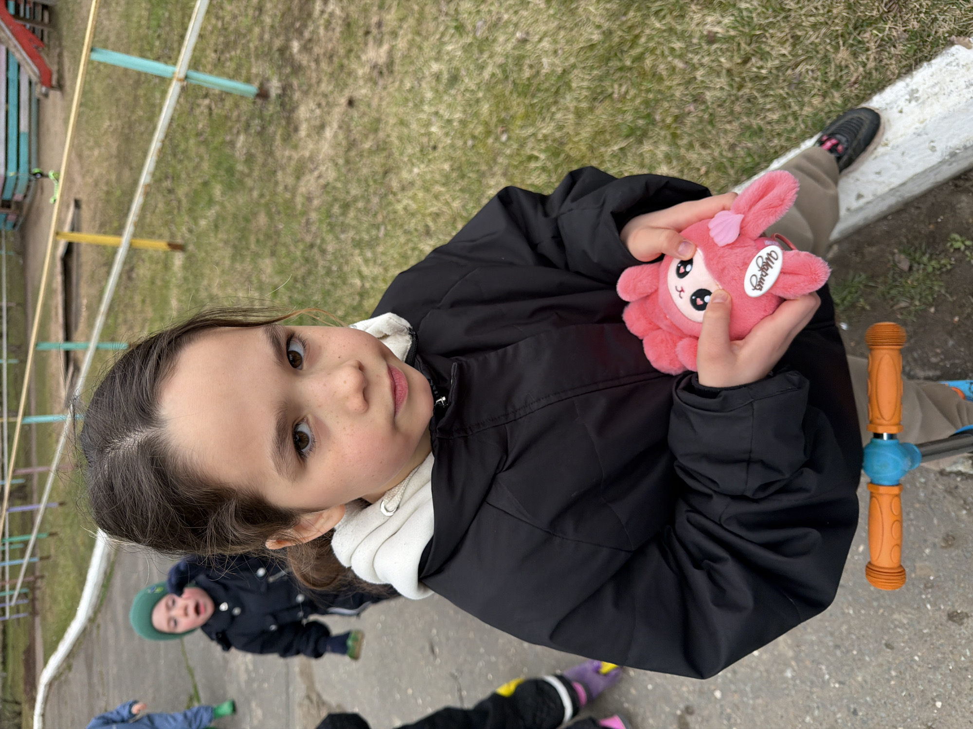 This image captures a young girl, estimated to be between 8 and 12 years old, looking directly at the camera with a calm, friendly expression and a slight smile. She has dark brown hair, light brown eyes, and noticeable freckles across her nose and cheeks. She is wearing a black puffer jacket over a white hooded sweatshirt, indicating cool weather. In her hands, she holds a pink plush toy resembling a bunny or a similar creature, featuring large eyes and a white tag with the text "Ugglys" or "Uggly's" clearly visible.
The scene unfolds in an outdoor playground or park setting in Chaikovsky, Russia. The ground is a mix of patchy, early-growth or dormant grass and bare earth. In the background, metal playground equipment, including bars painted in blue, yellow, and red, is partially visible. A concrete path or curb runs along the right side of the frame. Part of an orange and blue handlebar, likely from a scooter or balance bike, is also seen on the right.
In the lower left background, another younger child, dressed in a dark blue jacket and a green hat, is visible, looking upwards with an open mouth, suggesting they are in the midst of play or conversation. The legs and colorful shoes (purple and yellow) of a third child are partially seen at the very bottom. The activity taking place appears to be casual outdoor play amongst children during the daytime in cool weather, consistent with early spring or late autumn in the region.