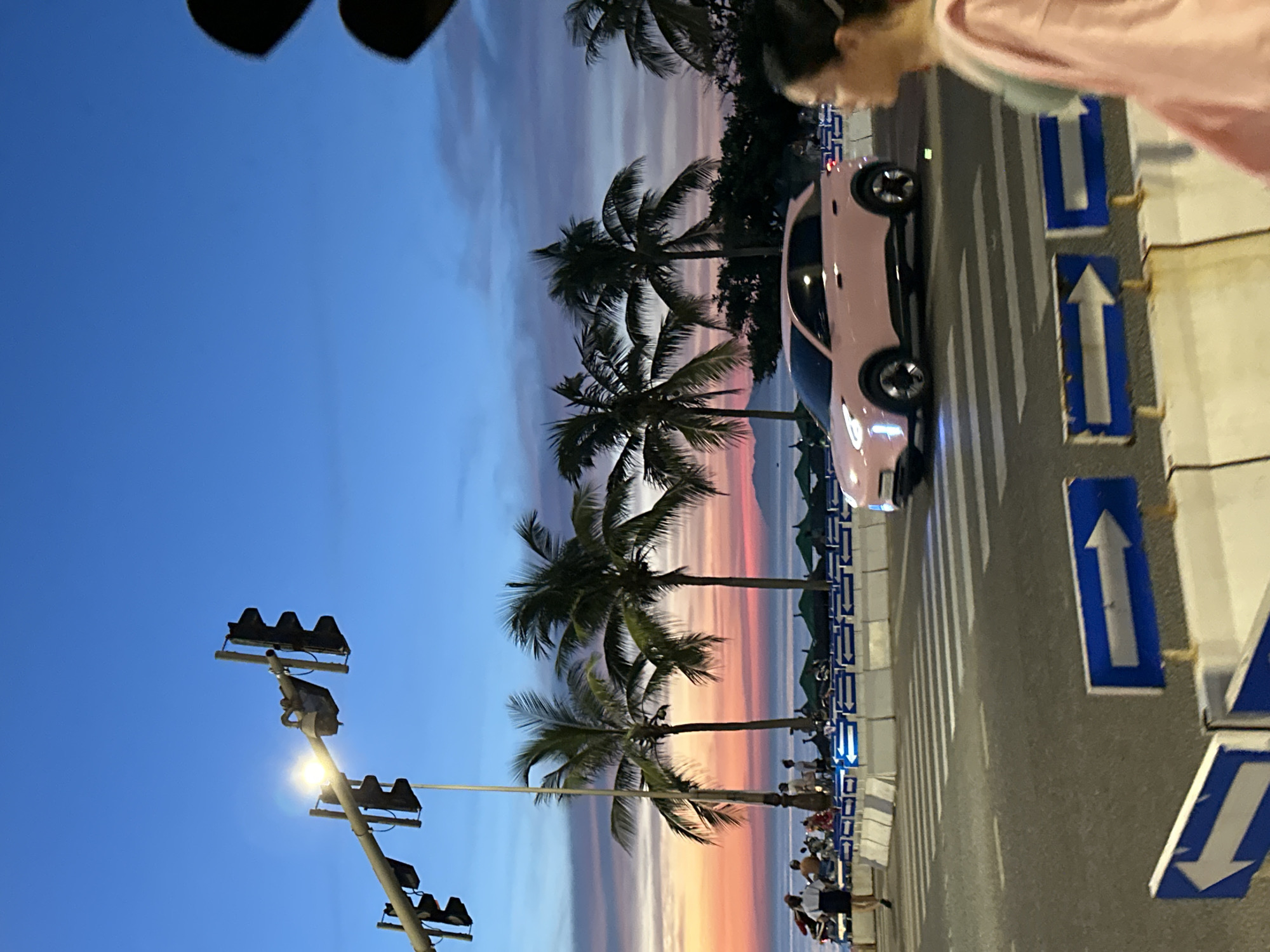 The image captures a street scene in Da Nang, Vietnam, during twilight. A pink electric car is driving across a pedestrian crossing on a paved road. On the right, a woman with dark hair, wearing a pink garment over a green top, is walking away from the camera. In the background, palm trees line a beach, with umbrellas set up on the sand and a body of water visible. A traffic light structure is prominent in the upper left quadrant of the image, against a clear blue and orange gradient sky. Several motorcycles and people are gathered near the beach area. White concrete barriers with blue directional arrows are placed along the side of the road.
